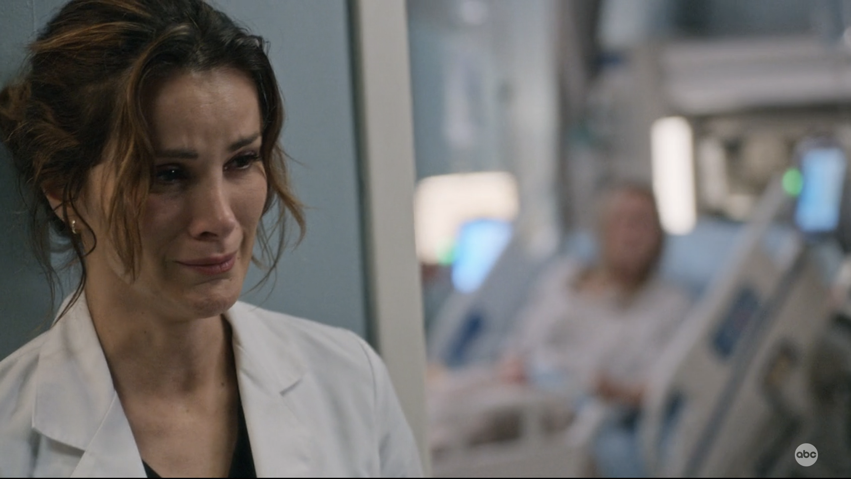 Carina cries in the foreground with Maya in a hospital bed blurred behind her 