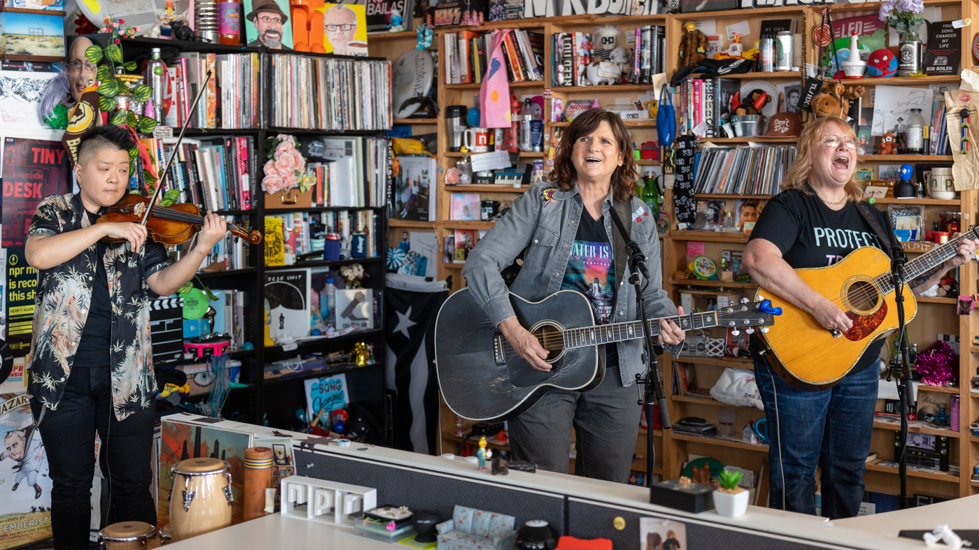 The Indigo Girls perform in a cramped library space with many books. They seem happy while singing.