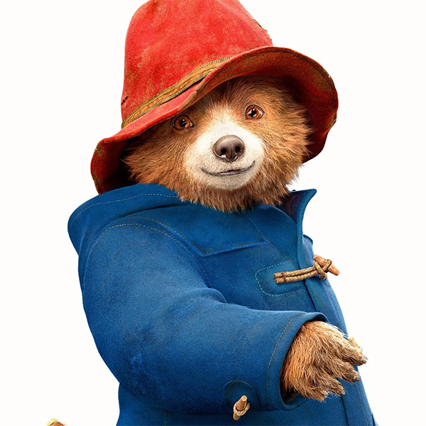 Paddington in his trademark blue pea coat and red hat. He's smiling.