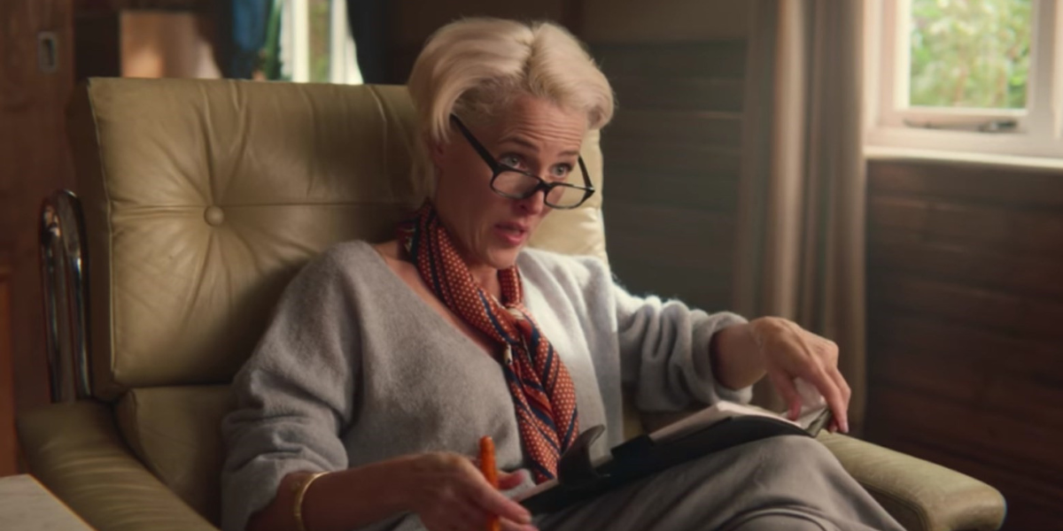 Gillian Anderson sits in an armchair on Sex Education, wearing a tiny orange scarf and a gray cashmere sweater, looking down eyeglasses