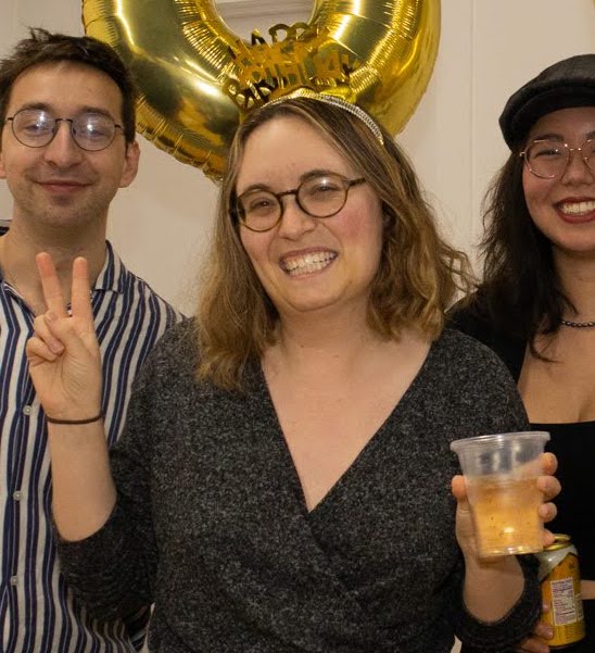 Anya smiling with a crown that says "Happy Birthday" and holding a golden drink. Anya is a white woman with shoulder length light brown hair and glasses