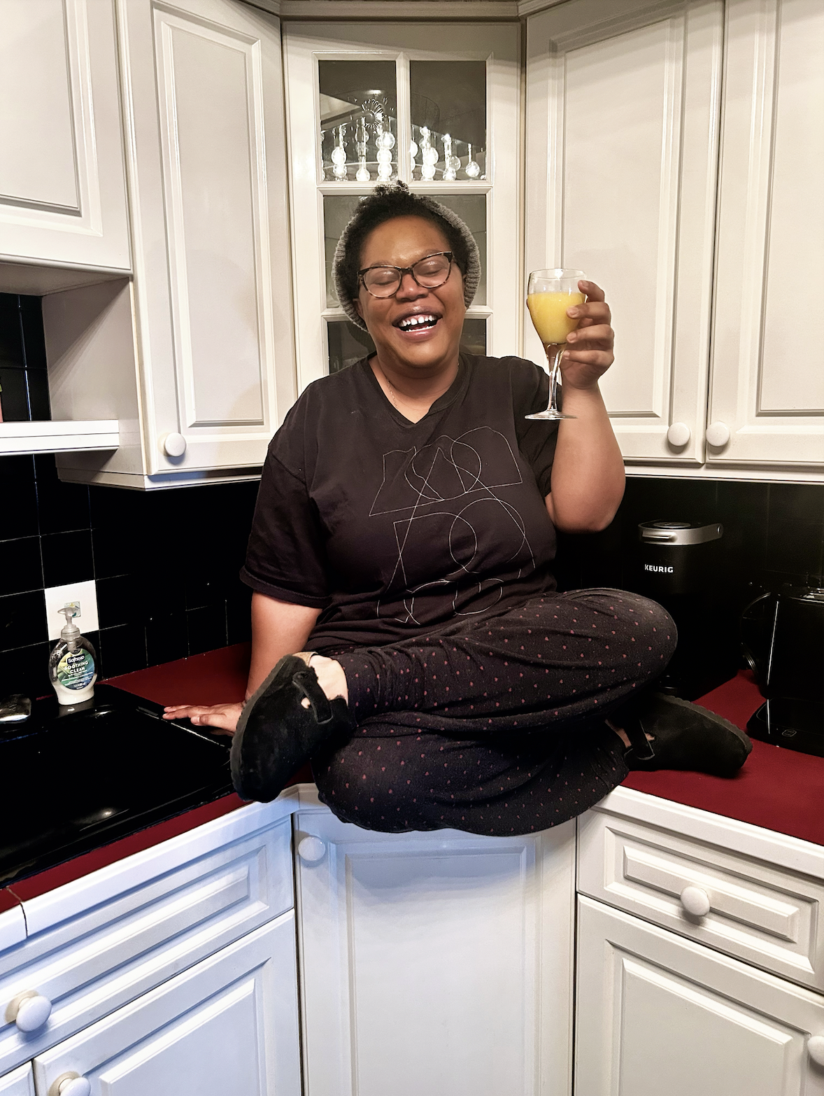 Carmen, a Black woman, sits on a kitchen counter, legs crossed, wearing a you do you tee, casual pants and birkenstocks. she is holding orange juice in a wine glass probably a mimosa and smiling