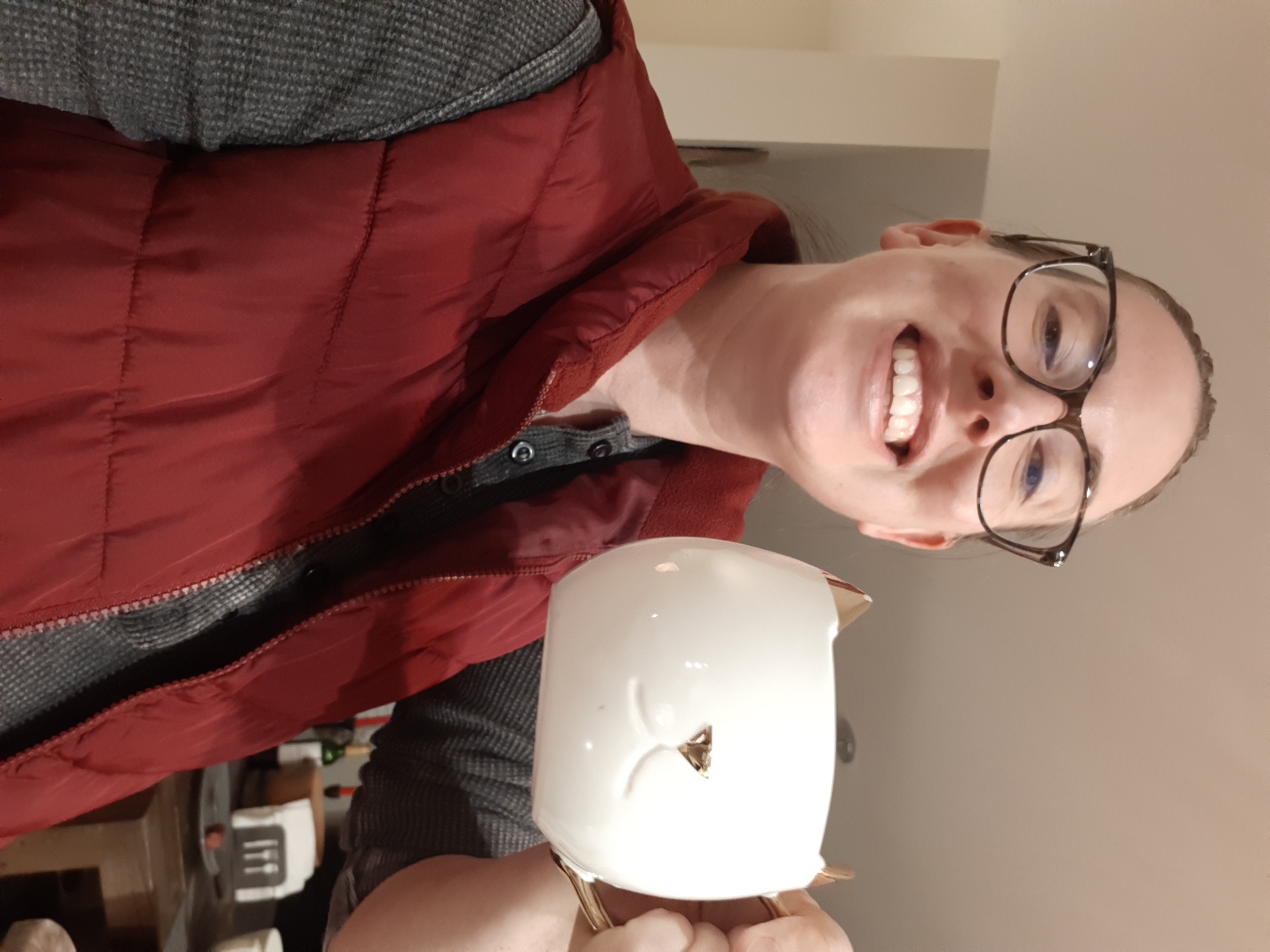casey is a white woman with glasses who is holding a mug in the shape of a cat's face up to the camera - she is wearing glasses