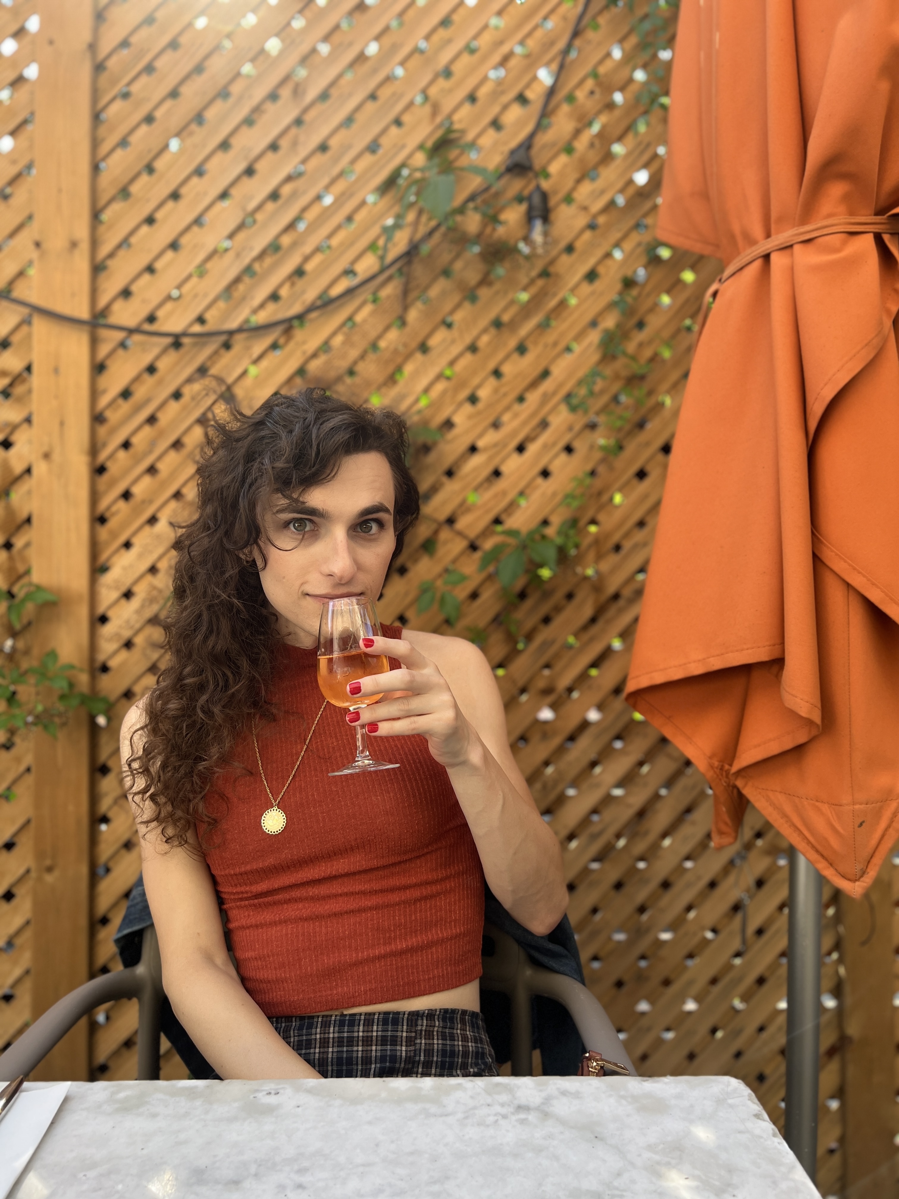 Drew a white woman with long curly hair is wearing a plaid skirt maybe and a rust colored sleeveless top. she outside sipping on either orange wine or an aperol spritz and looking at the camera