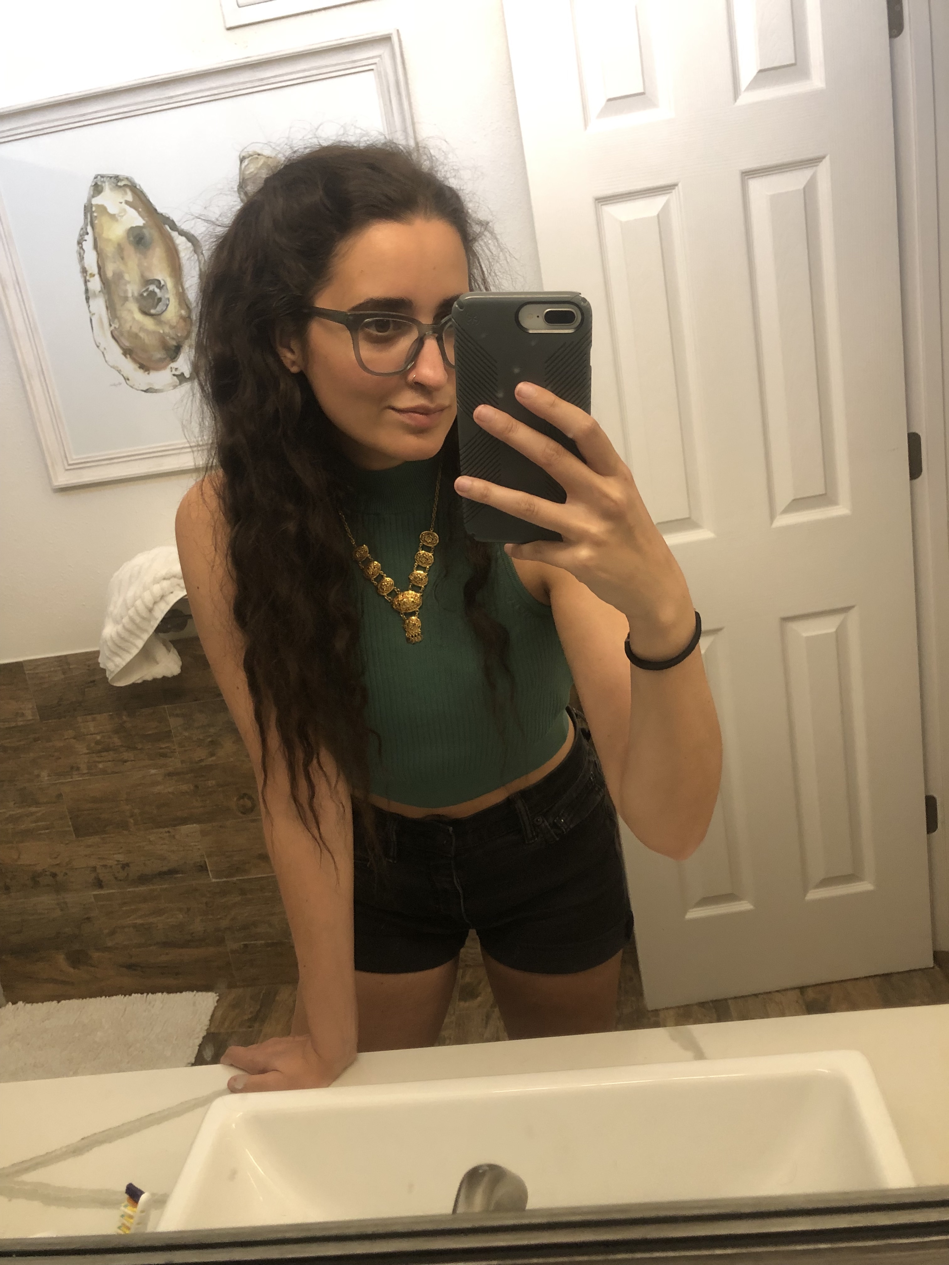 Kayla Kumari Upadhyaya wears a cropped green knit tank and a gold necklace over black jean shorts. Her hair is done in two small side partial-pigtails. She is wearing blue glasses.