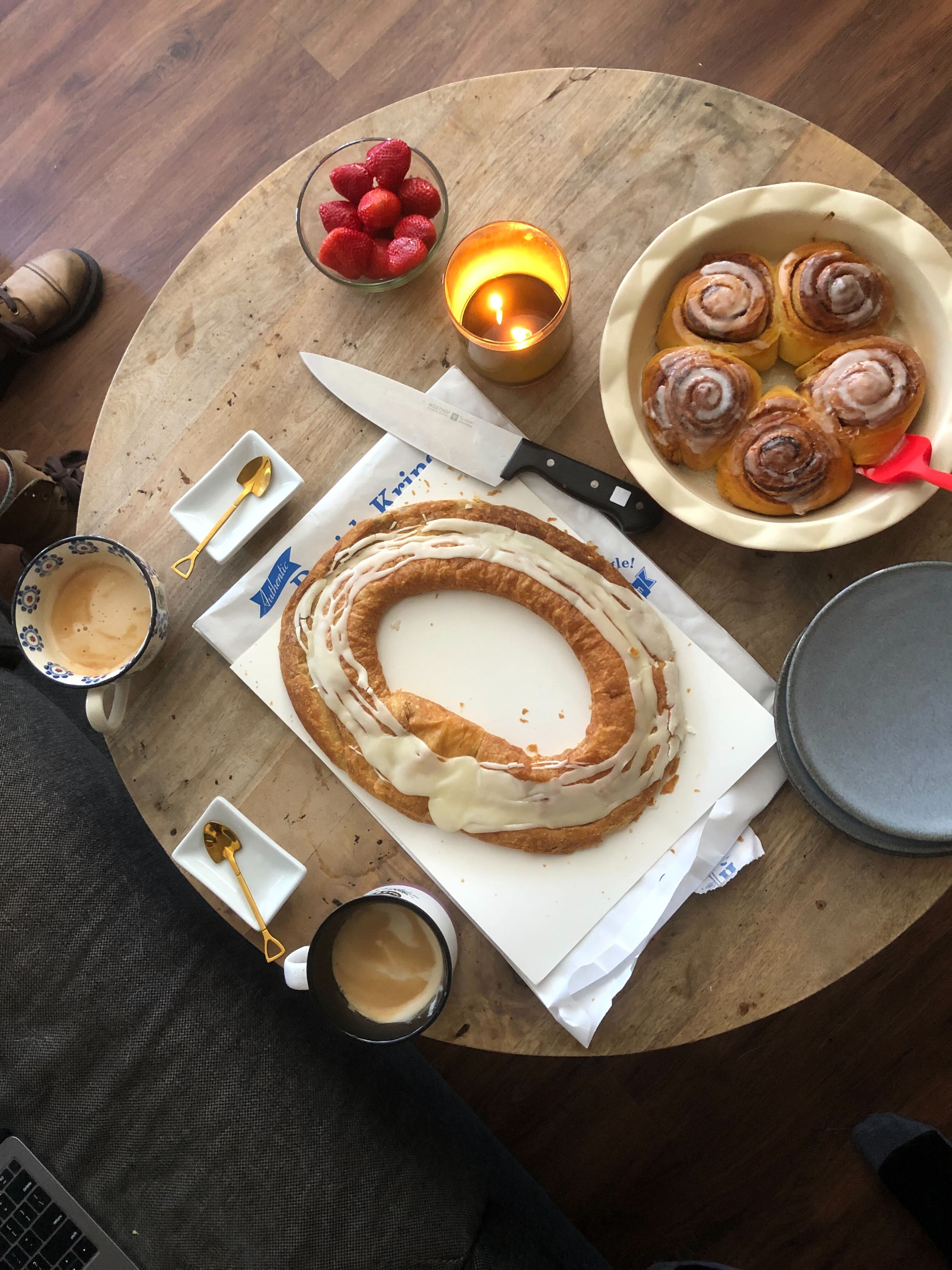 a table with cinnamon rolls, strawberries, lattes, tiny golden shovels, and a kringle with a knife
