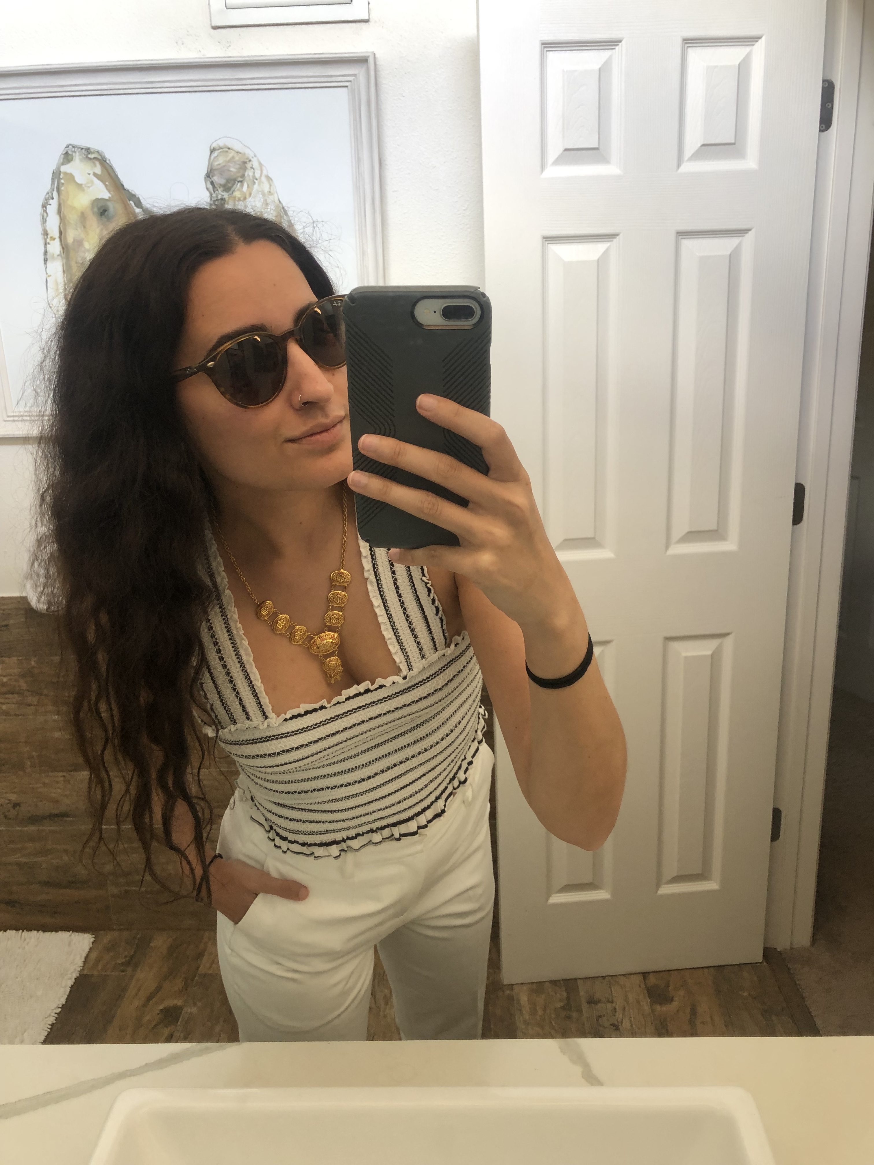 Kayla Kumari Upadhyaya is wearing sunglasses and a white an blue striped stretchy cropped tank with a gold necklace as well as tailored white pants. Her hair is down and parted in the middle.