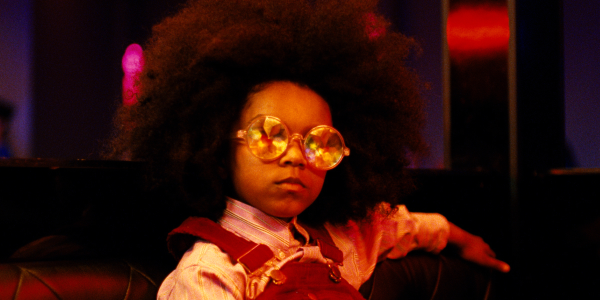 Sally Dramé as Vicky wears kaleidoscope glasses and looks toward the camera.