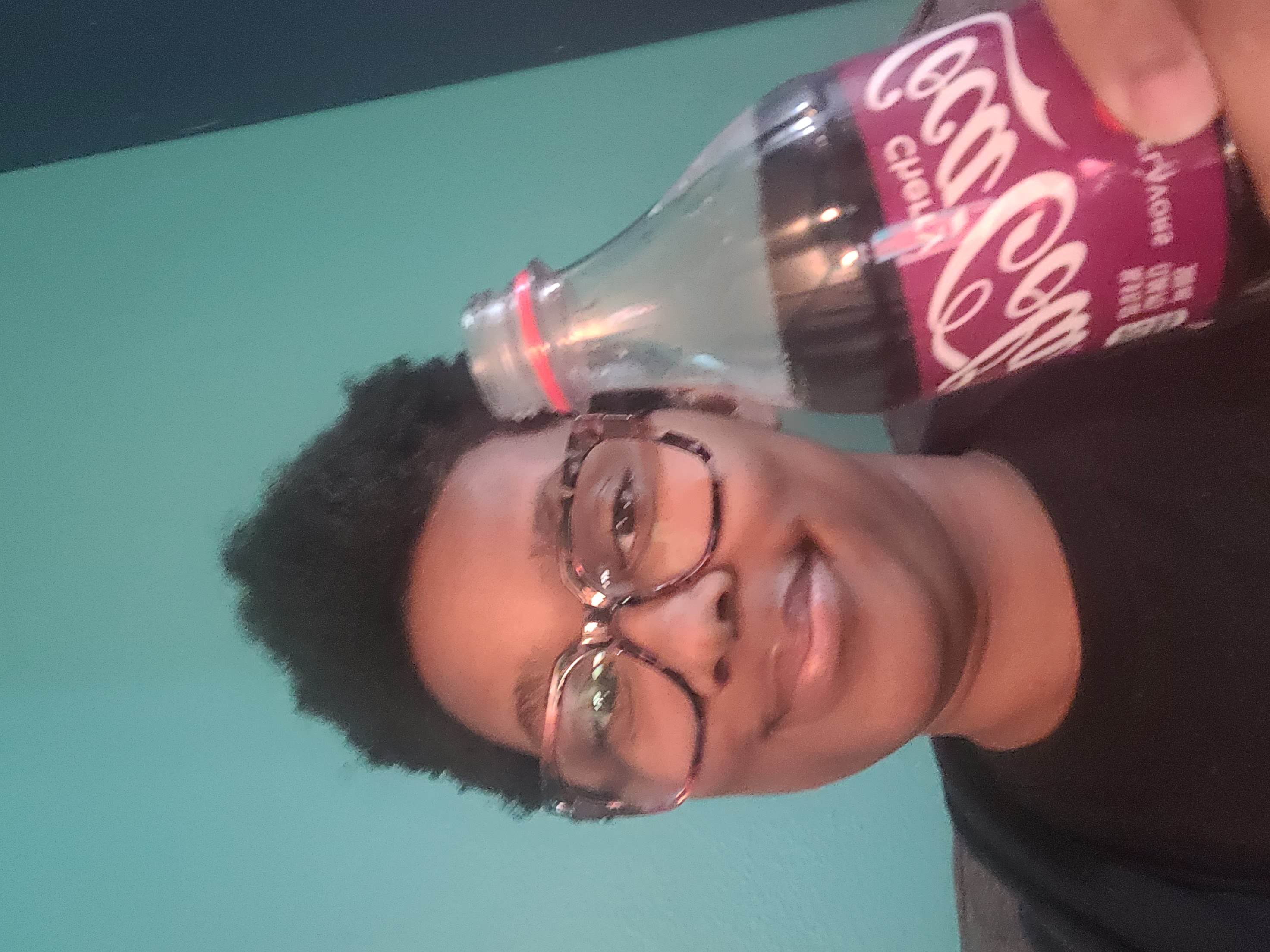 Sa'iyda, a Black woman wearing glasses holds up a cherry coke to the camera and smiles