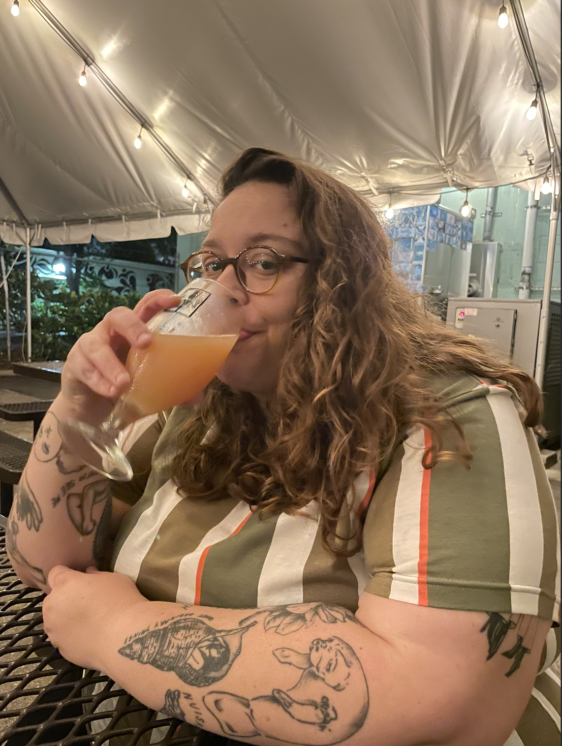 Stef, a white nonbinary person, drinks a beverage and looks at the camera. they are wearing a vertically striped top and have curly longer hair and glasses