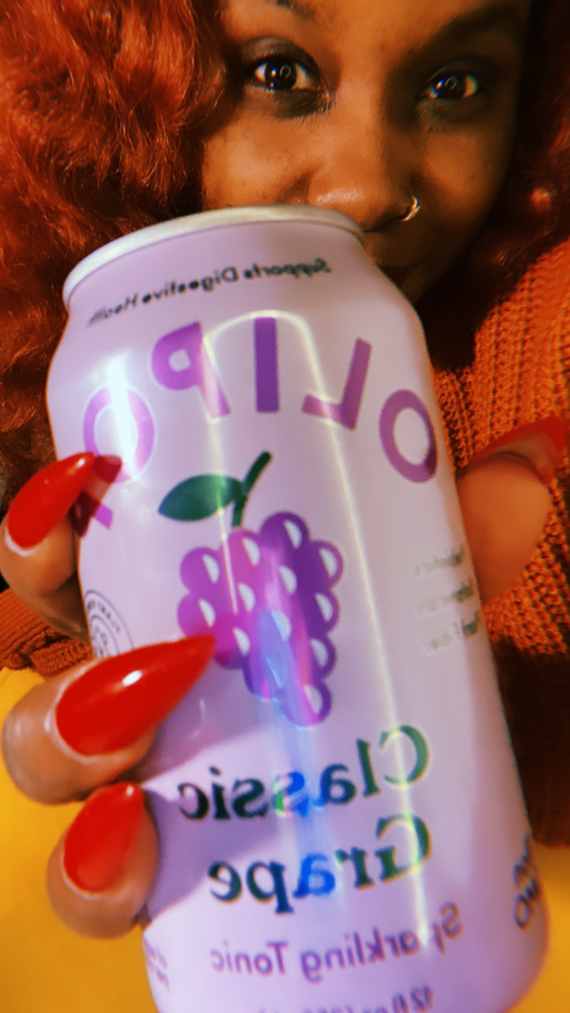 Shelli a Black woman with bright orange hair wearing an orange top with orange nails holds a can of classic grape olipop up to the camera