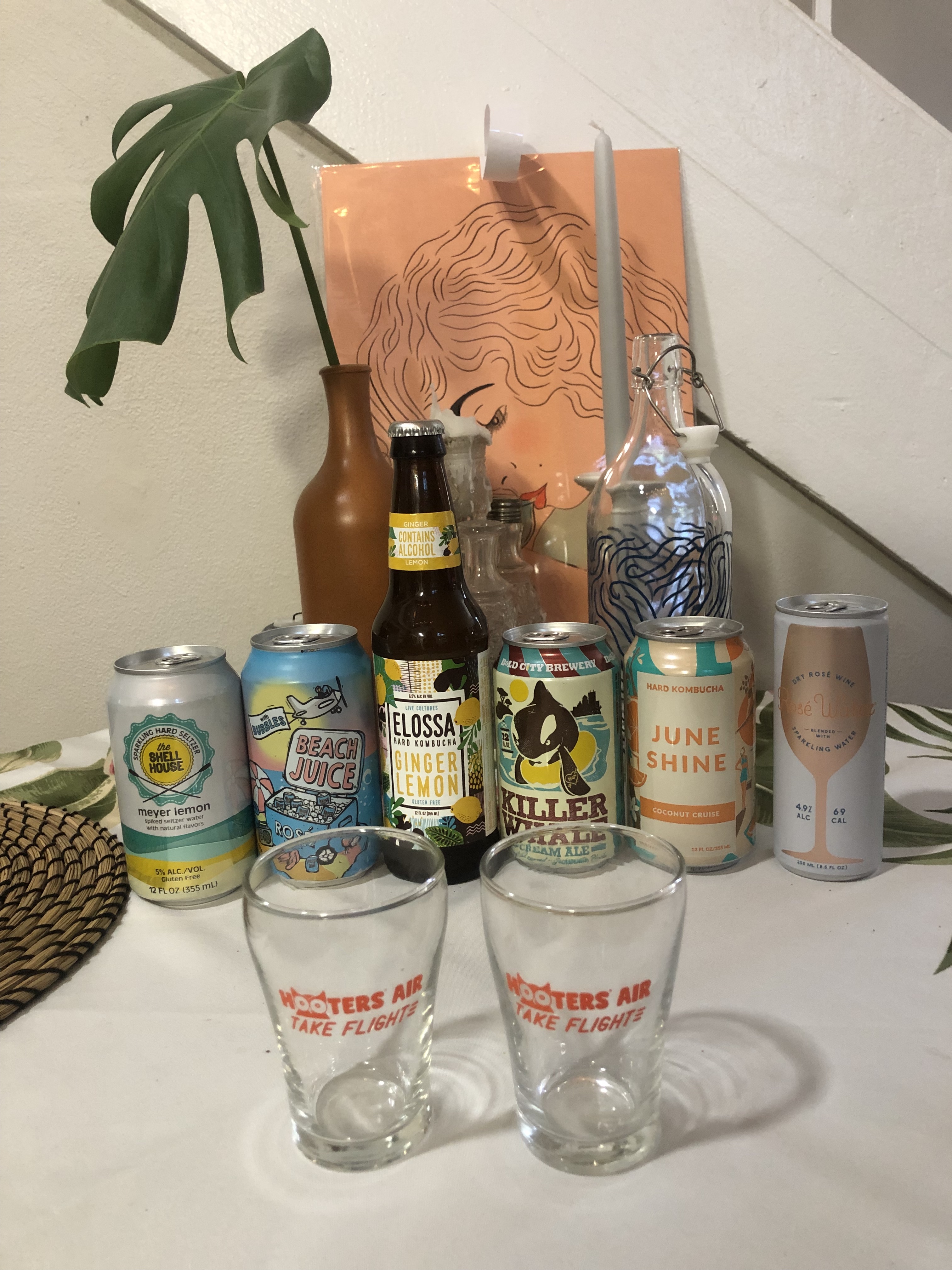 two cans of sparkling rose, two cans of hard kombucha, a can of hard seltzer, and a can of cream ale. there are two small Hooters glasses in front of the drinks.