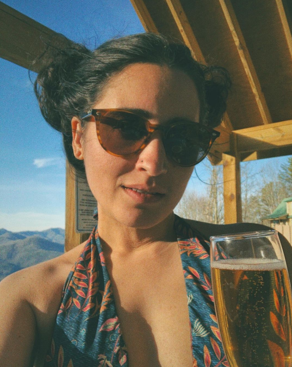 Kayla is a South Asian woman who here has her long dark hair up in two buns and is wearing sunglasses and a patterned bathing suit and holding up a glass of sparkling wine to the camera.