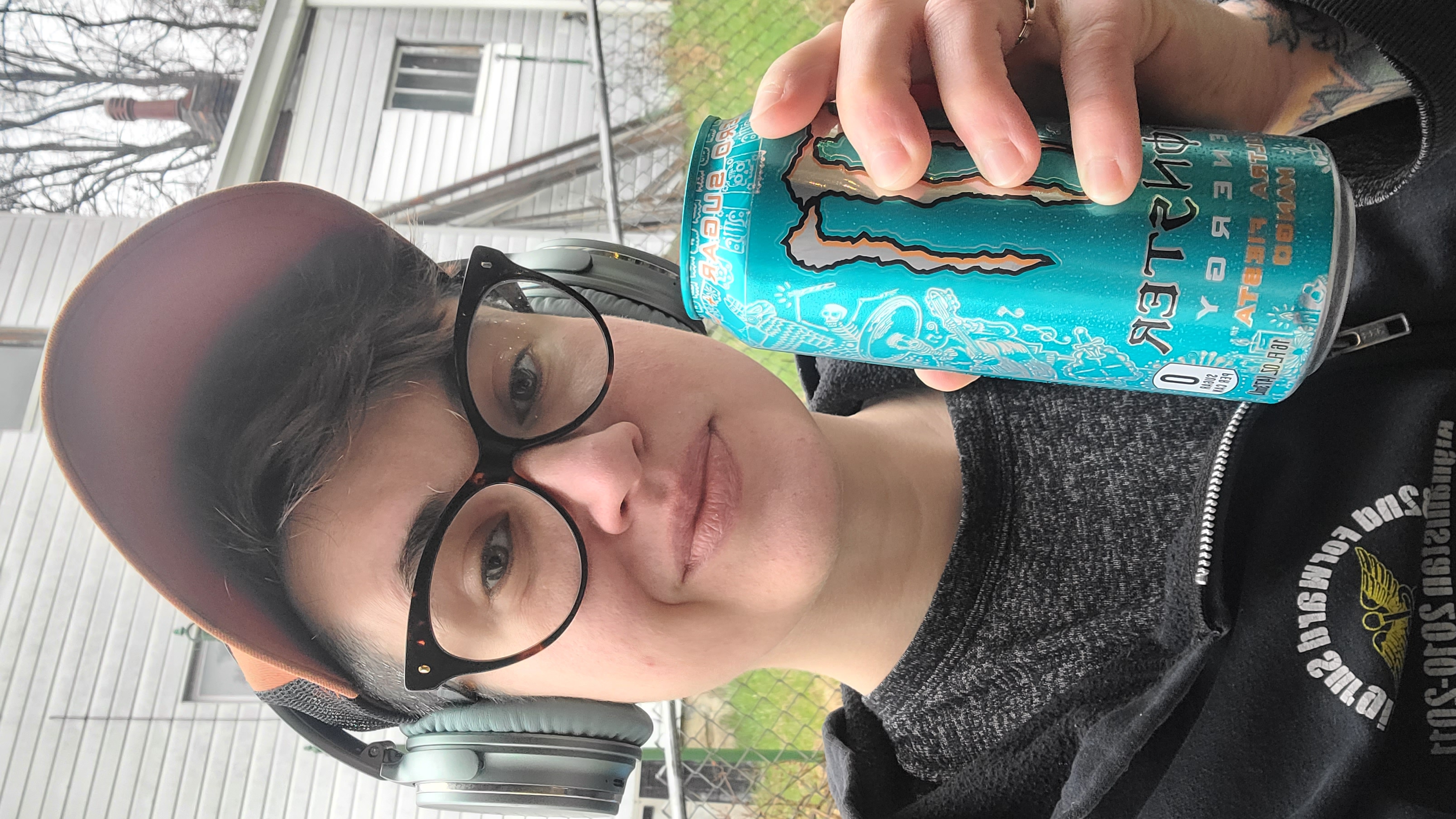 Nico is a white genderqueer human who is here wearing a trucker hat, headphones, glasses and a black sweatshirt and is holding up a monster can to the camera. they have shaved sides and wavy brown hair on top.