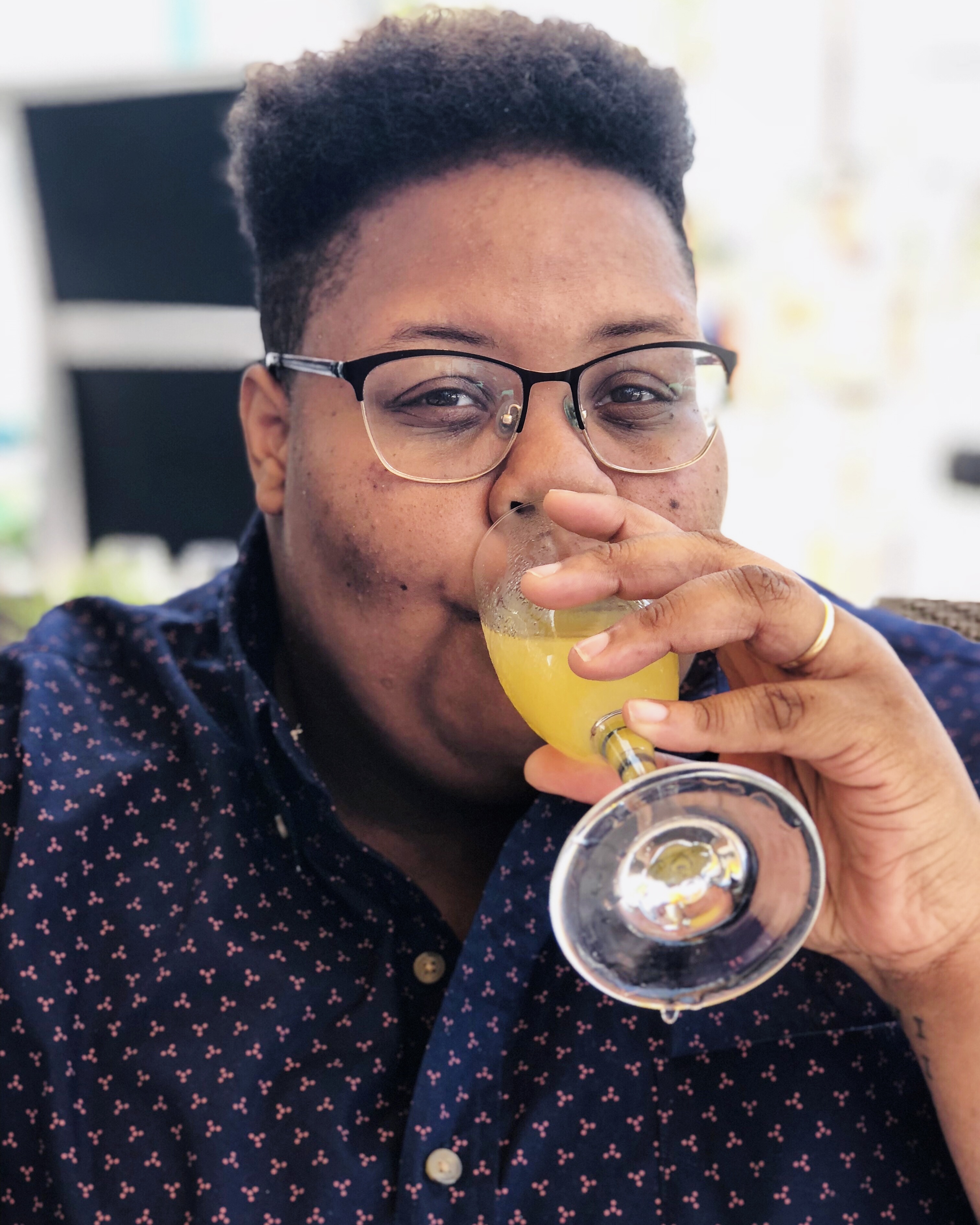 shea, a Black nonbinary human with short hair and glasses wearing a patterned button up smiles and drinks a mimosa 