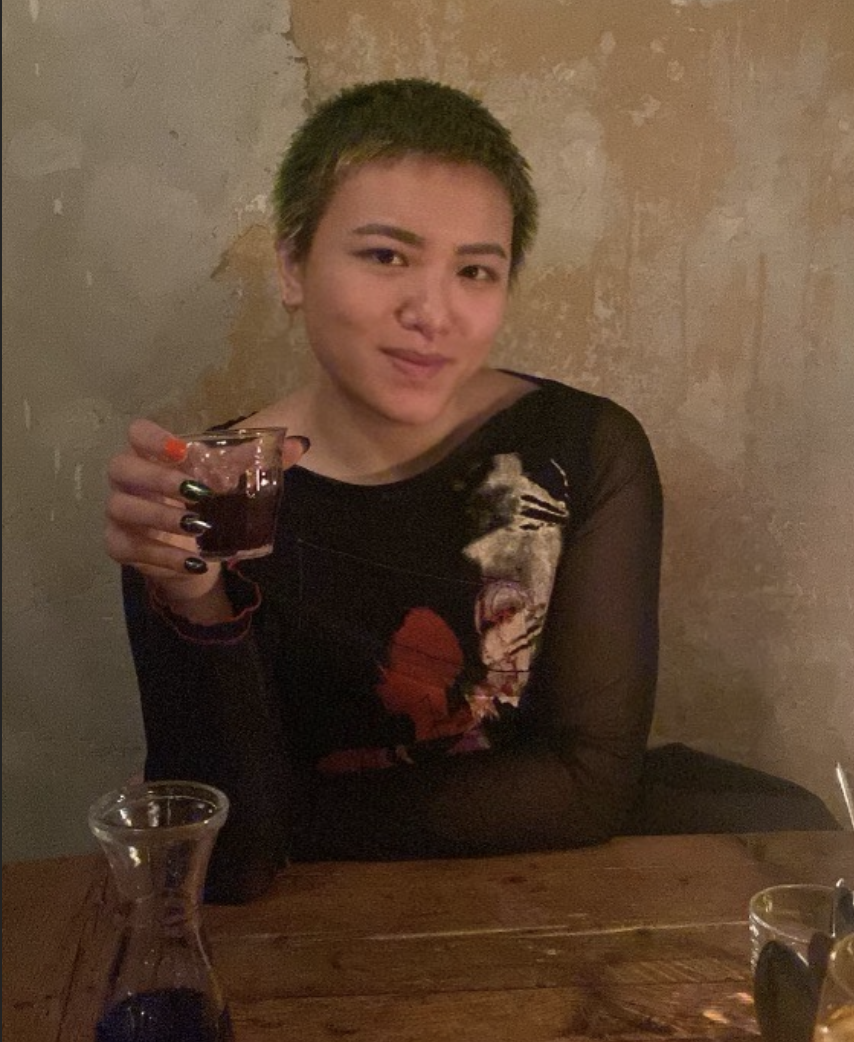 Viv, an Asian nonbinary person with short green hair is seated in a bar in Berlin holding a glass of red wine and smiling mischieviously. they are wearing an artsy black and abstractly patterned shirt.