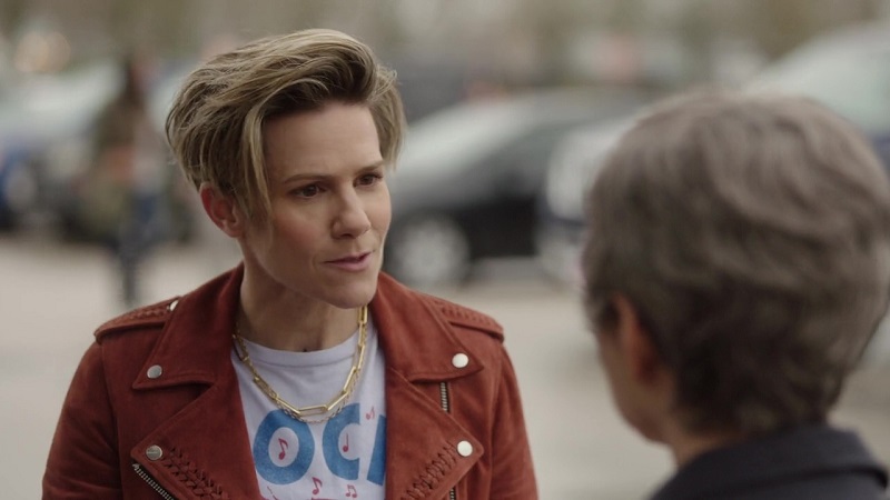 Greta is astonished when her future mother-in-law offers her money for her upcoming wedding. Greta is wearing a burnt orange corduroy jacket over a white t-shirt. Katherine's mom has her back to the audience.