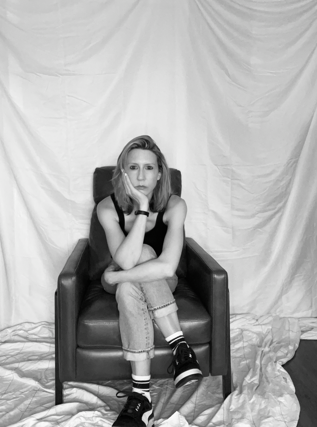 a black and white photo of riese, a white woman, with shoulder length blonde hair, sits cross legged on a chair with a background falling behind her.