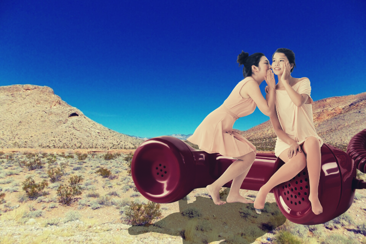 two women sit on a giant red landline phone, gossiping, with the desert as a background