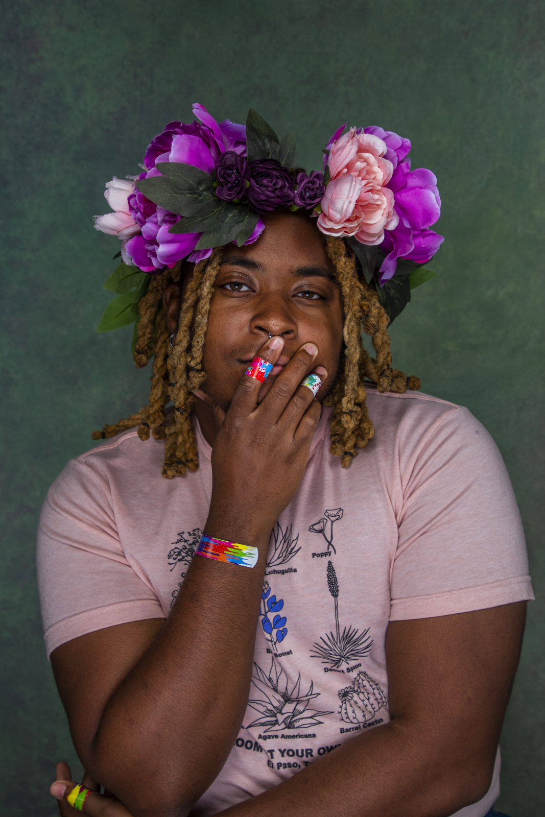 portrait of kb brookins, a black trans person with a flower crown, holding their face where multi colored band-aids can be seen on their hands and wrist. They are wearing a pink top and looking at the viewer. They have golden shoulder length twists.