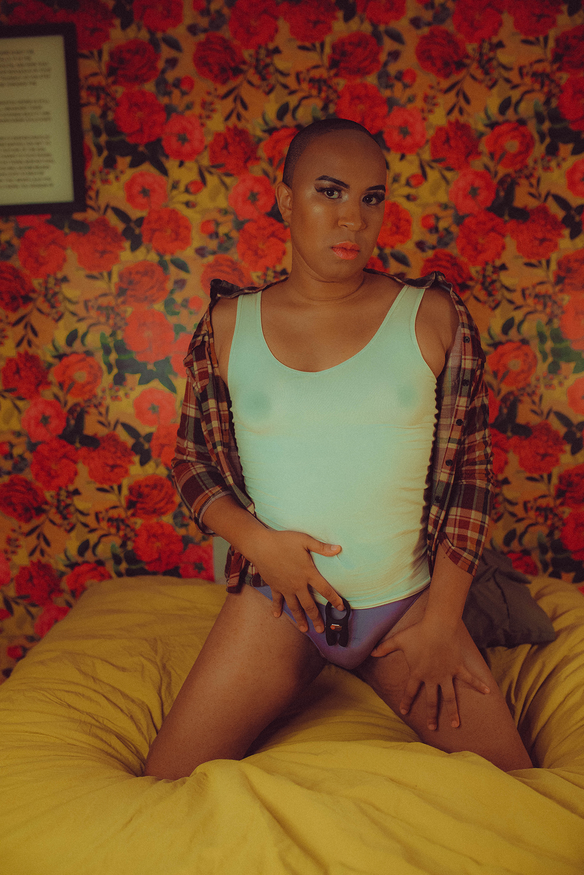 A Black person with a shaved head is kneeling on a yellow bedspread in front of floral wallpaper. They're wearing pink lipstick, dark eyeliner, a white tank top, purple underwear, and a multicolored flannel that's hanging off of their shoulders. They hold a black Fun Factory vibrating cock ring in one hand near their pelvis.