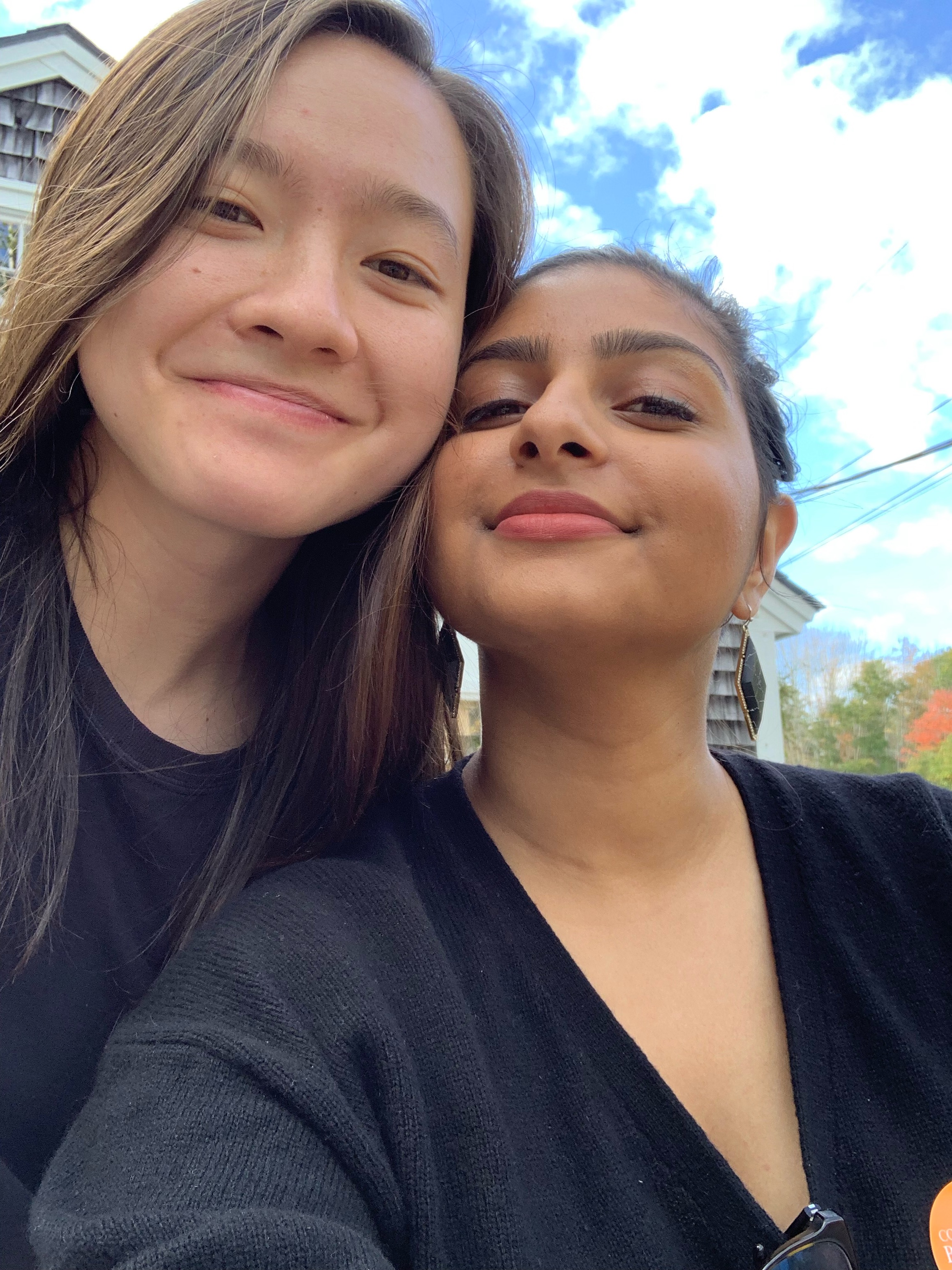 MR (left) and Ashni (right) are both smiling at the camera, wearing black shirts. MR's hair is long, Ashni's is in a bun. Ashni has on black geometric earrings.