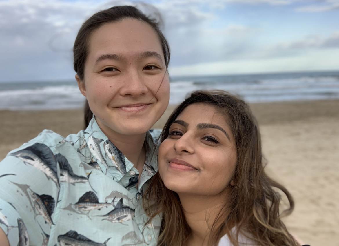 MR (left) and Ashni (right) on a beach, with the water behind them. Both people have their hair a little ruffled by the wind and are wearing blue button-downs.