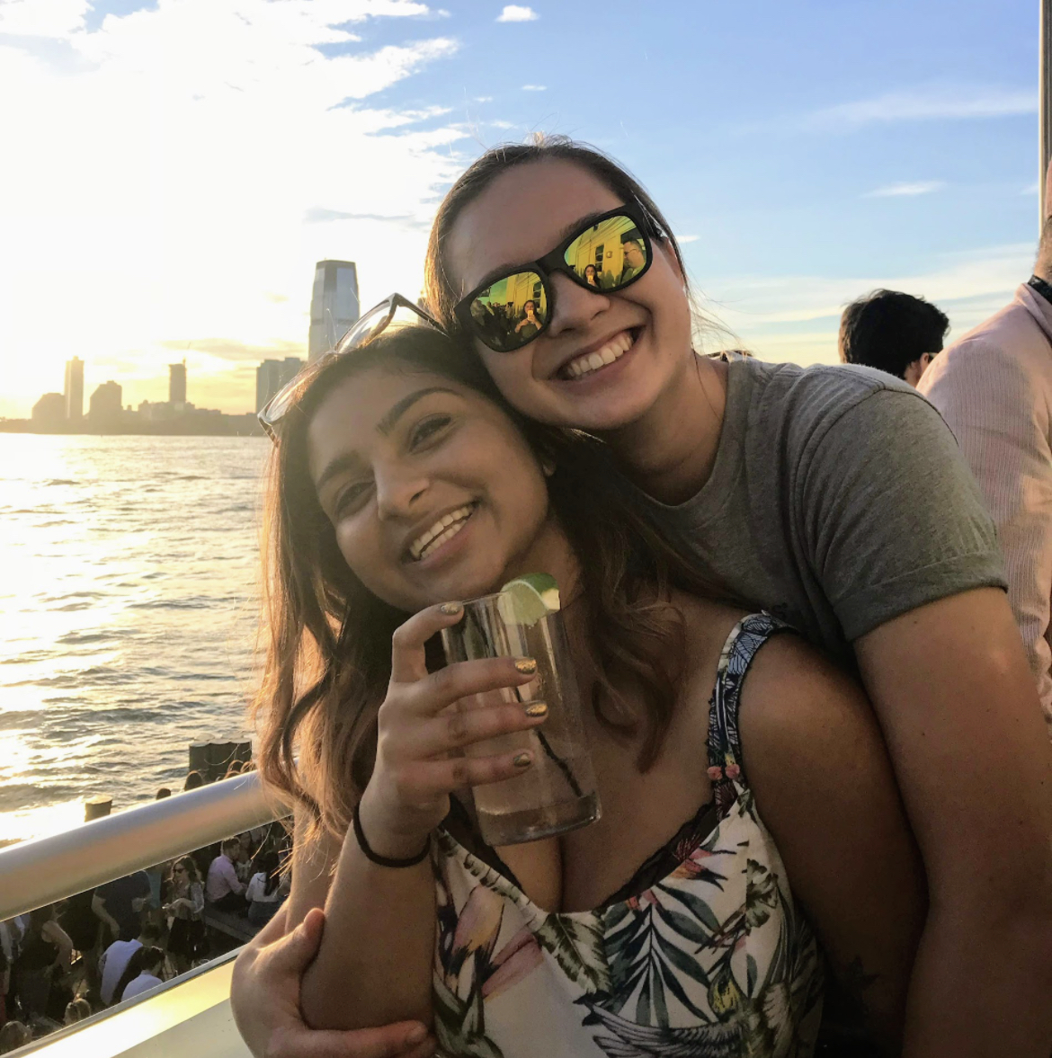 MR (they/them) and Ashni (they/she) posing for the camera. MR's arms are around Ashni's, and they are on a boat on the water. Ashni is holding a beverage.