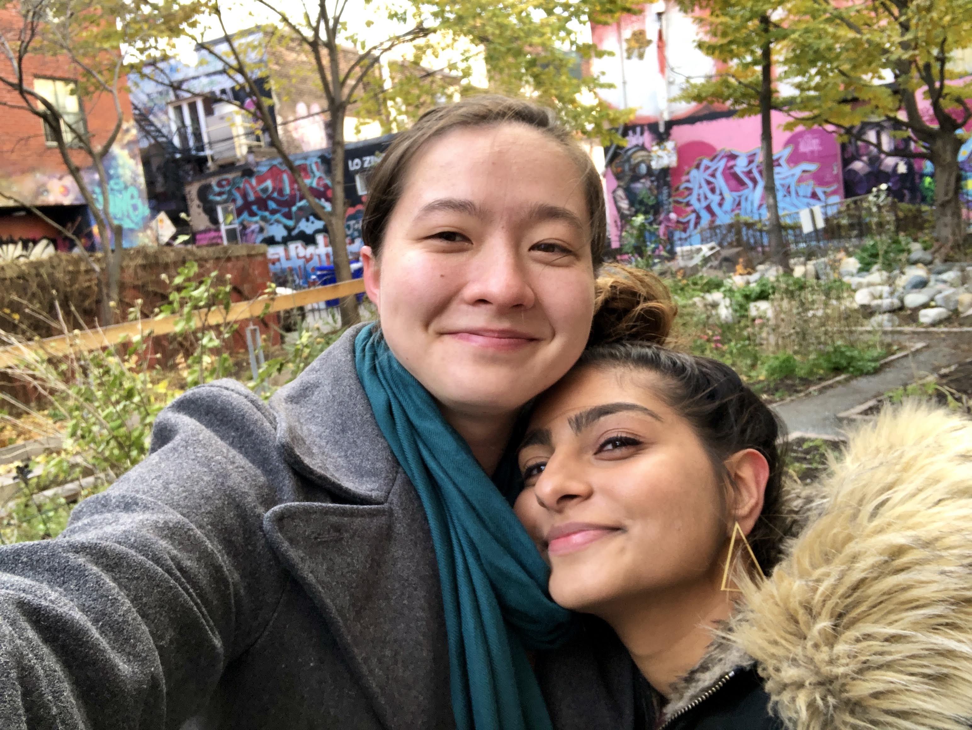 MR (left) and Ashni (right) are looking up at the camera. Ashni has their hair in a bun and is wearing triangle earrings. MR is wearing a blue scarf and a gray wool jacket.