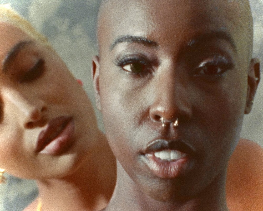 Two Black queer people holding each other while looking at the camera.