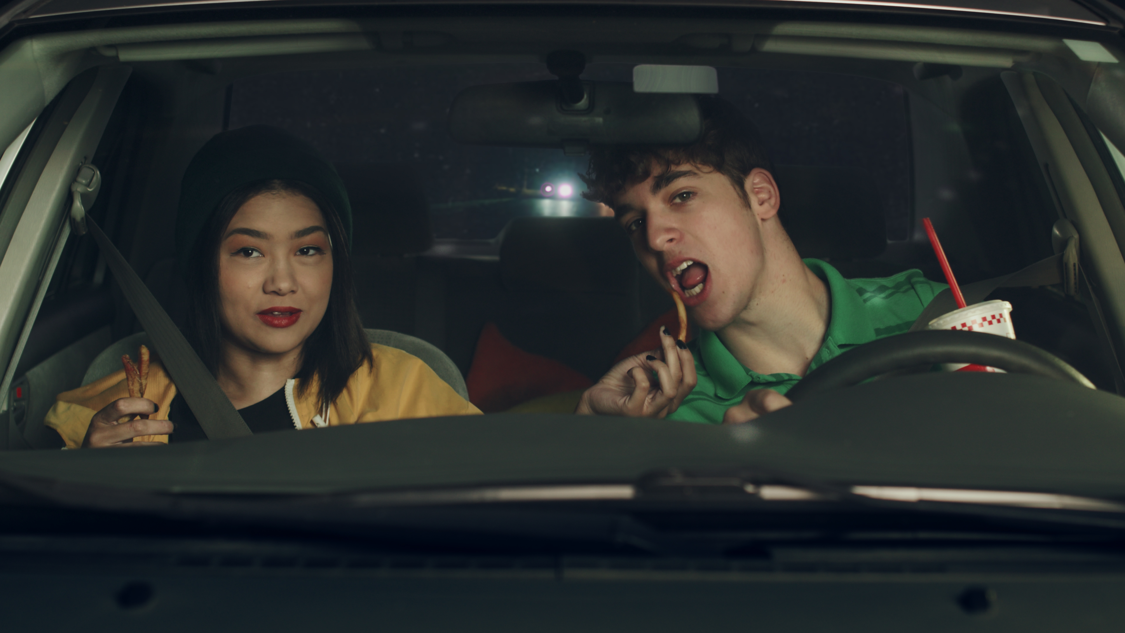 An Asian teen girl and a white teenage boy share a French fry in a car at night.