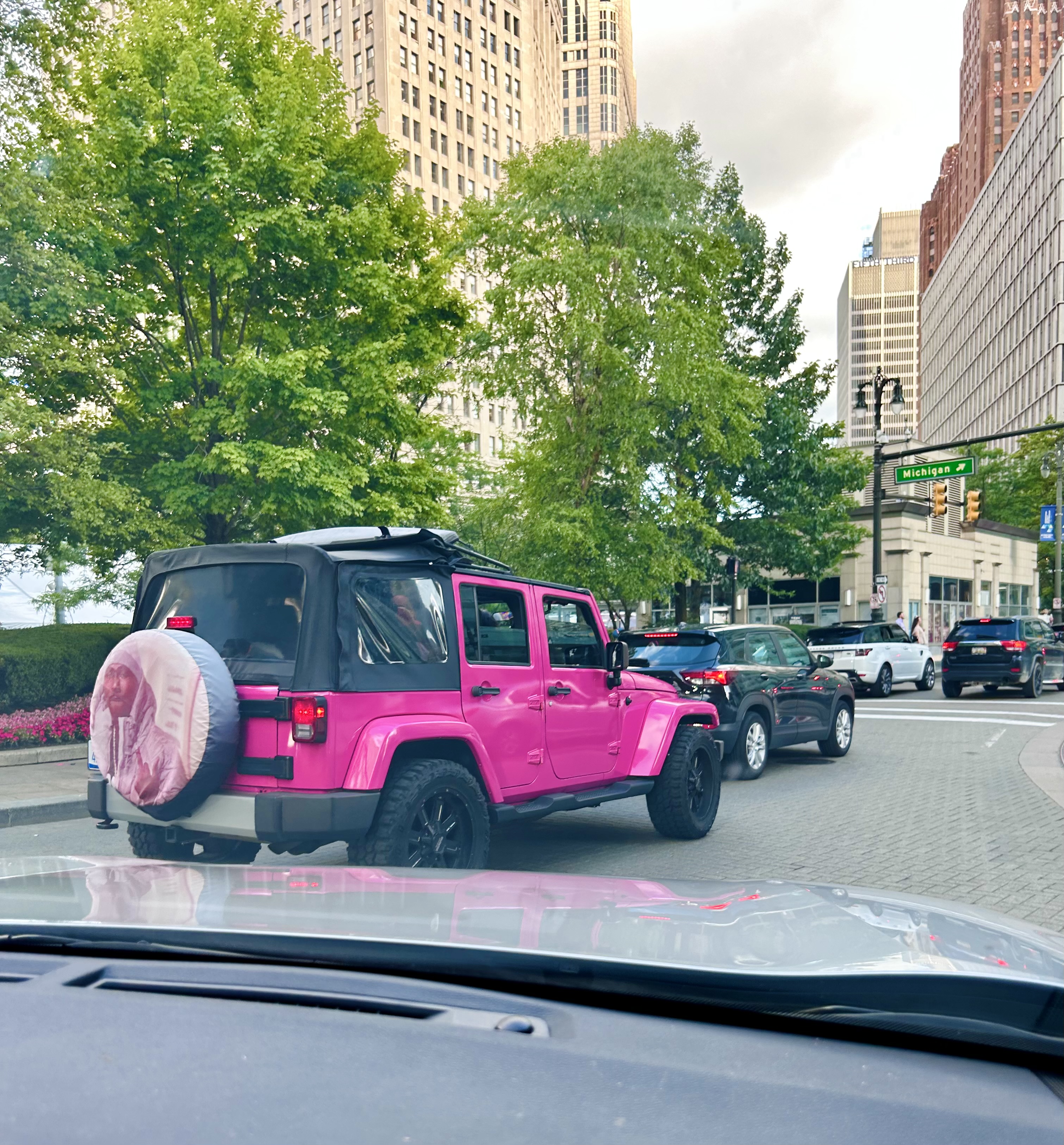 A hot pink jeep is driving around downtown Detroit, on the back wheel is a pink wheel cover with a rapper on it.