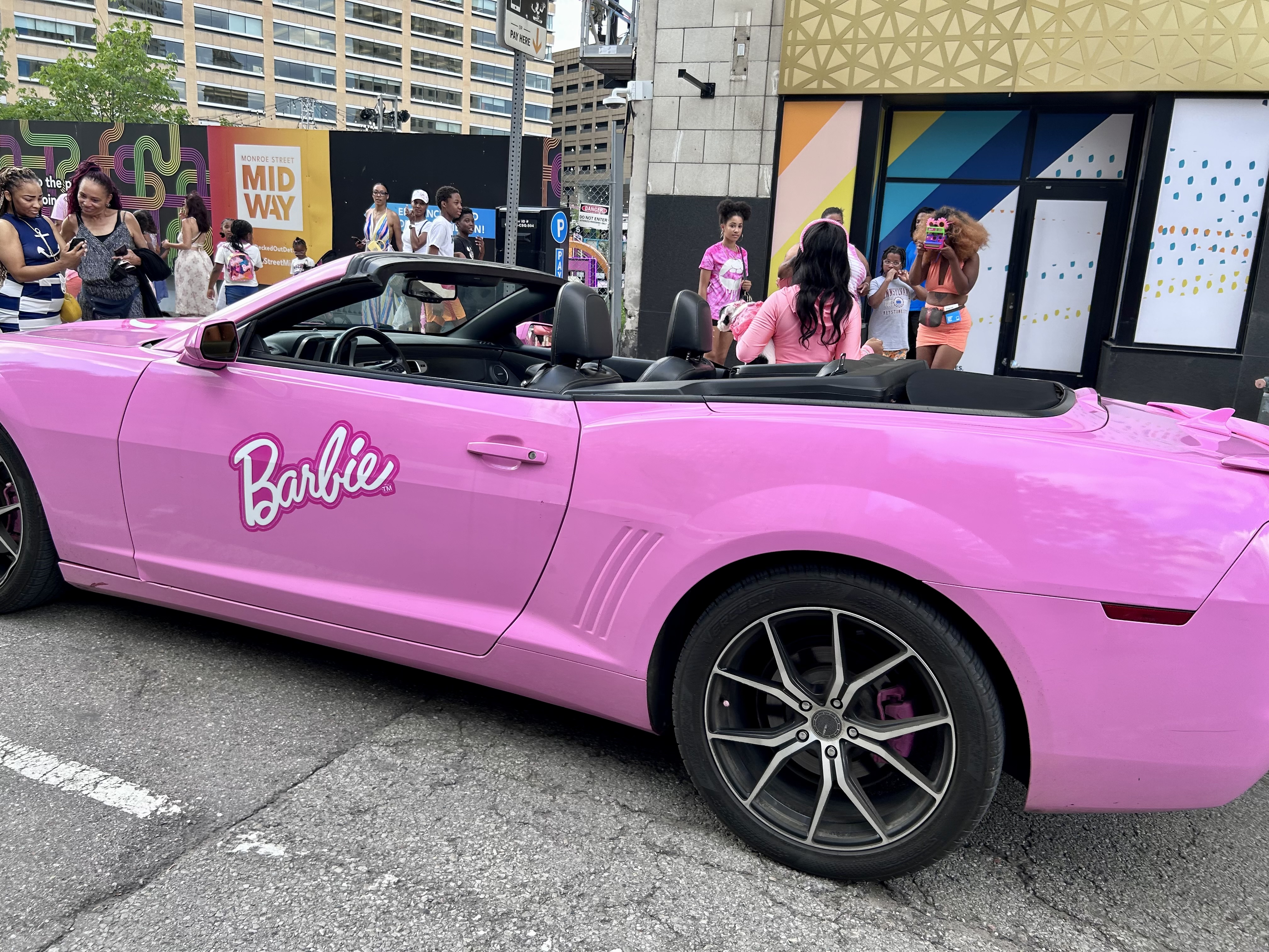 A pink corvette says "barbie" on the side in downtown Detroit, people are taking pictures beside it.