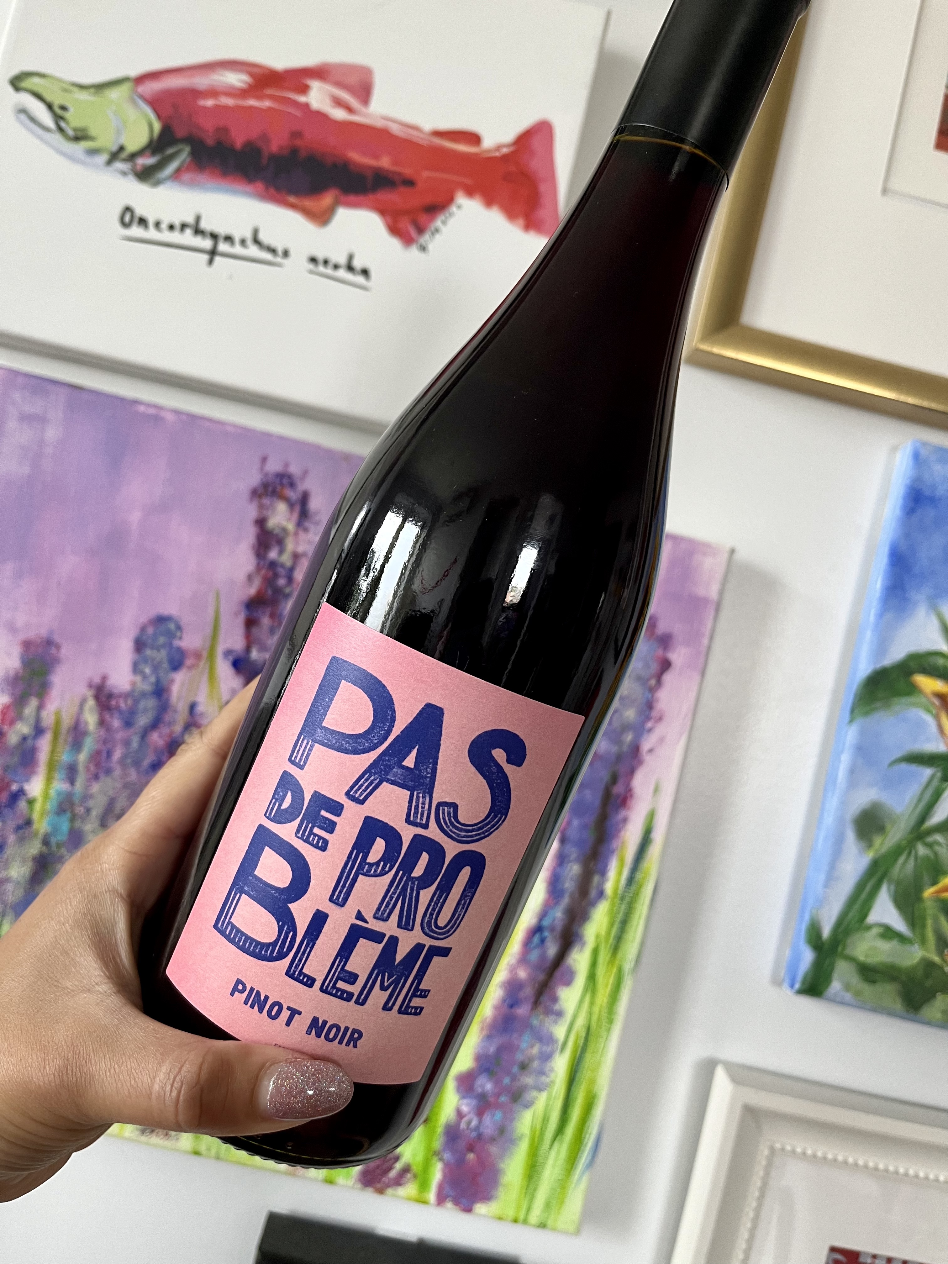A hand holding a dark bottle of red wine against a gallery wall.