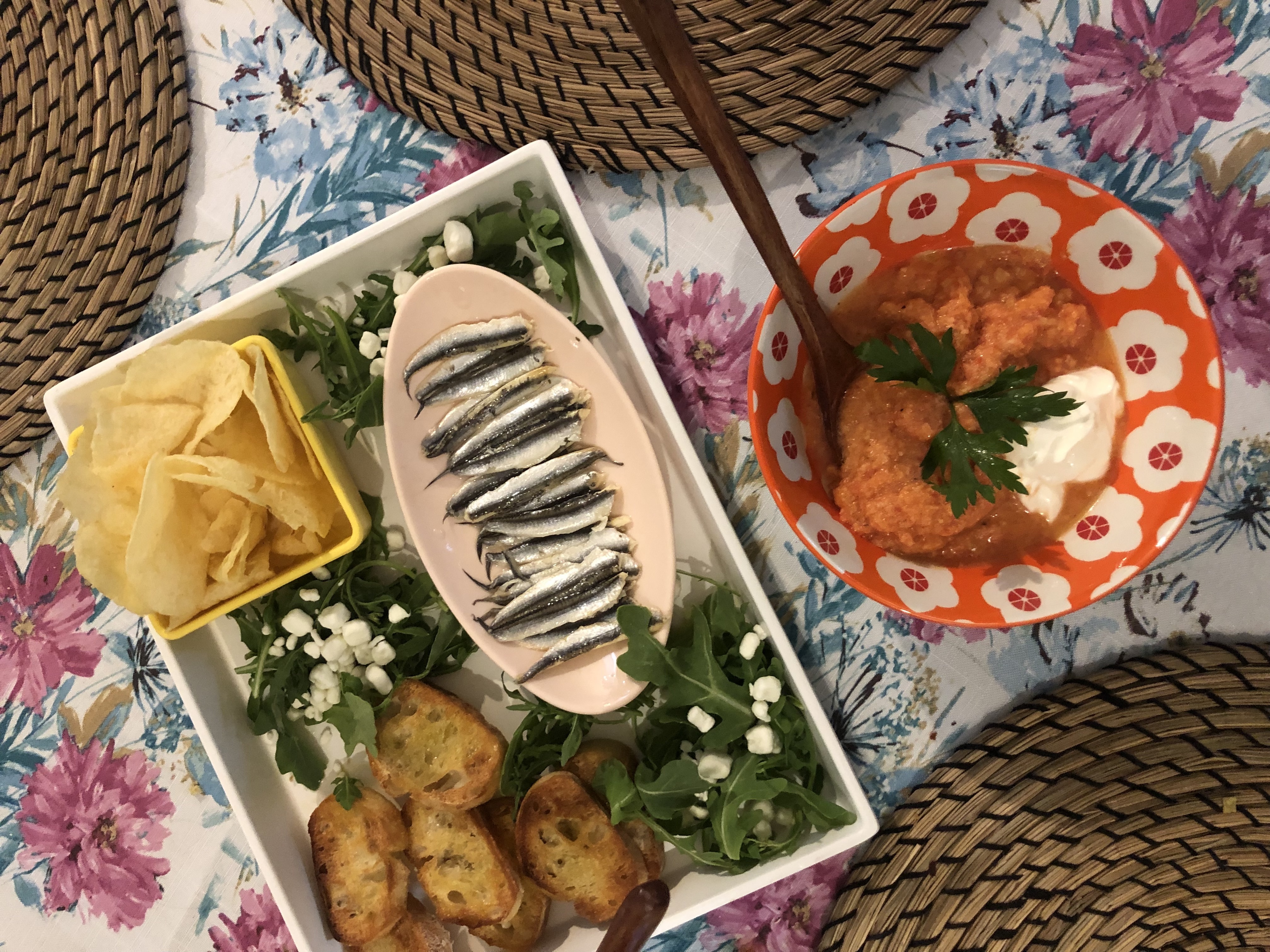 a bowl of gazpacho and a snack plate with arugula, goat cheese, potato chips, and crostini on it