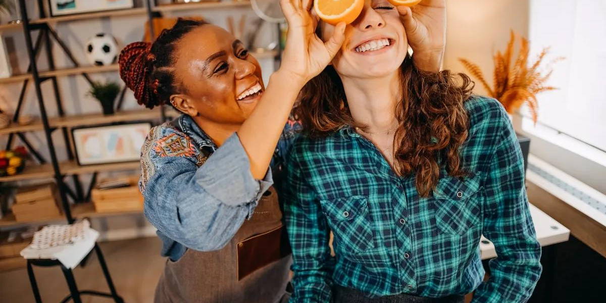 a woman holds orange halves in front of her girlfriend's eyeballs