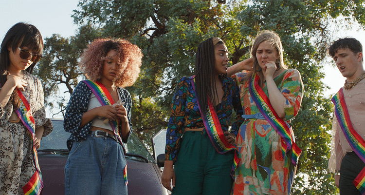the cast of Sissy stands in rainbow sashes, looking at something on a road