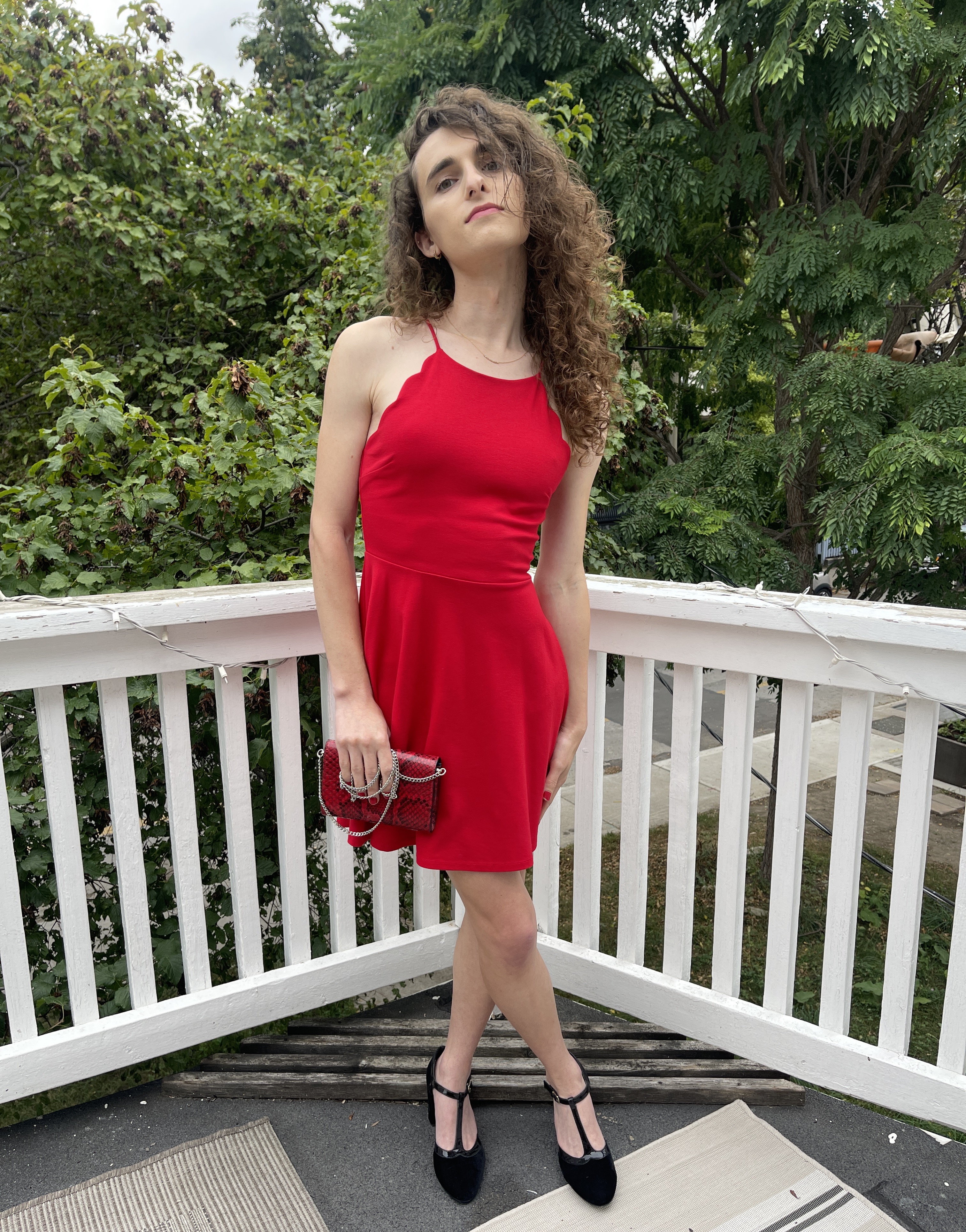 Drew poses on a balcony in a red dress. She clutches a red purse, is wearing black shoes, and has her head tilted up, her hair partially covering half her face.
