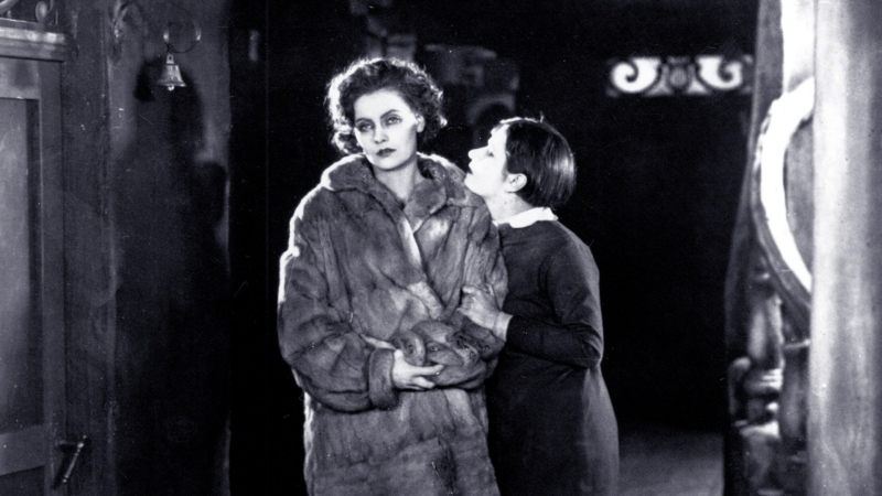 A young Greta Garbo in a fur coat looks forlorn as another woman grabs onto her arm.