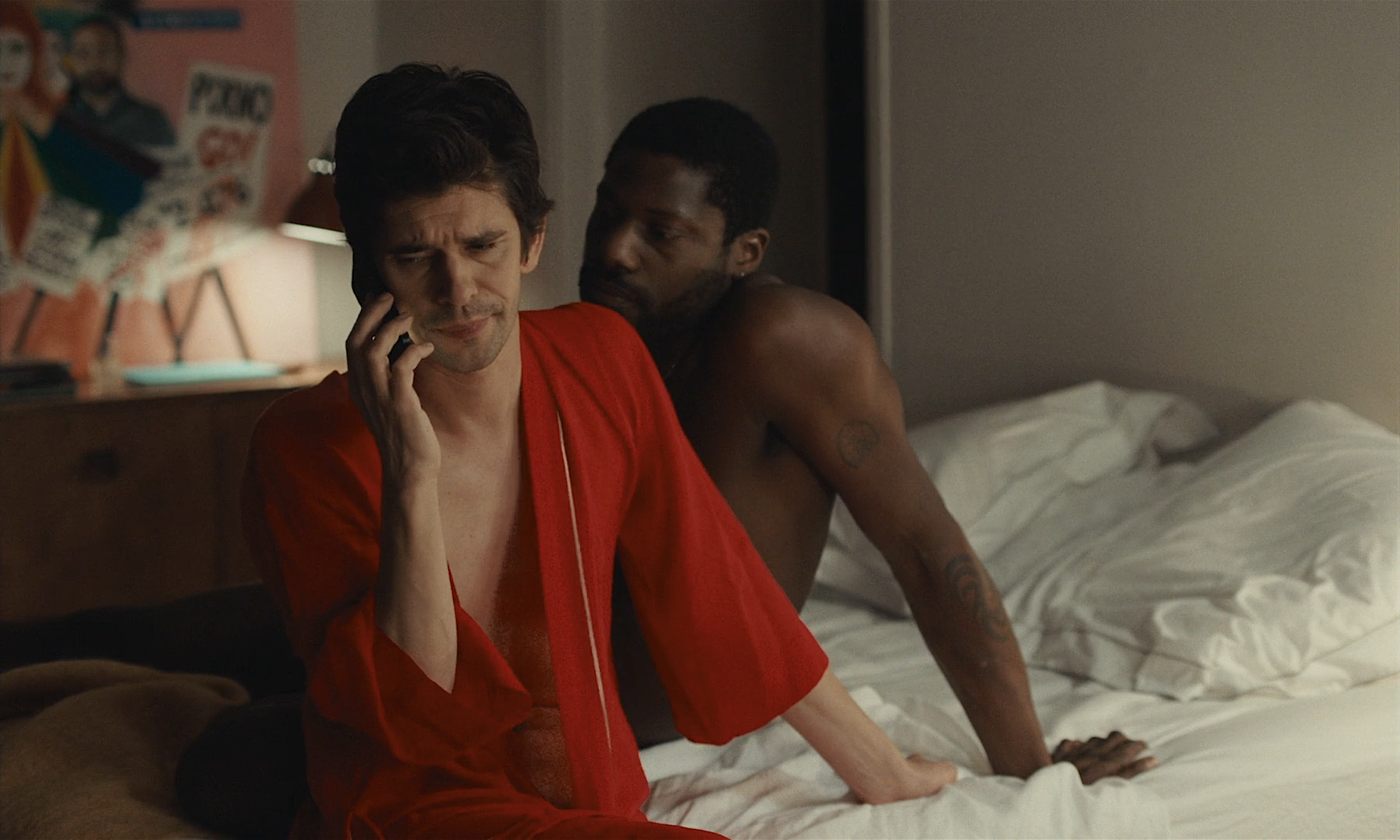 Ben Whishaw in an open red robe talks on the phone. A shirtless man sits up in bed besides him.
