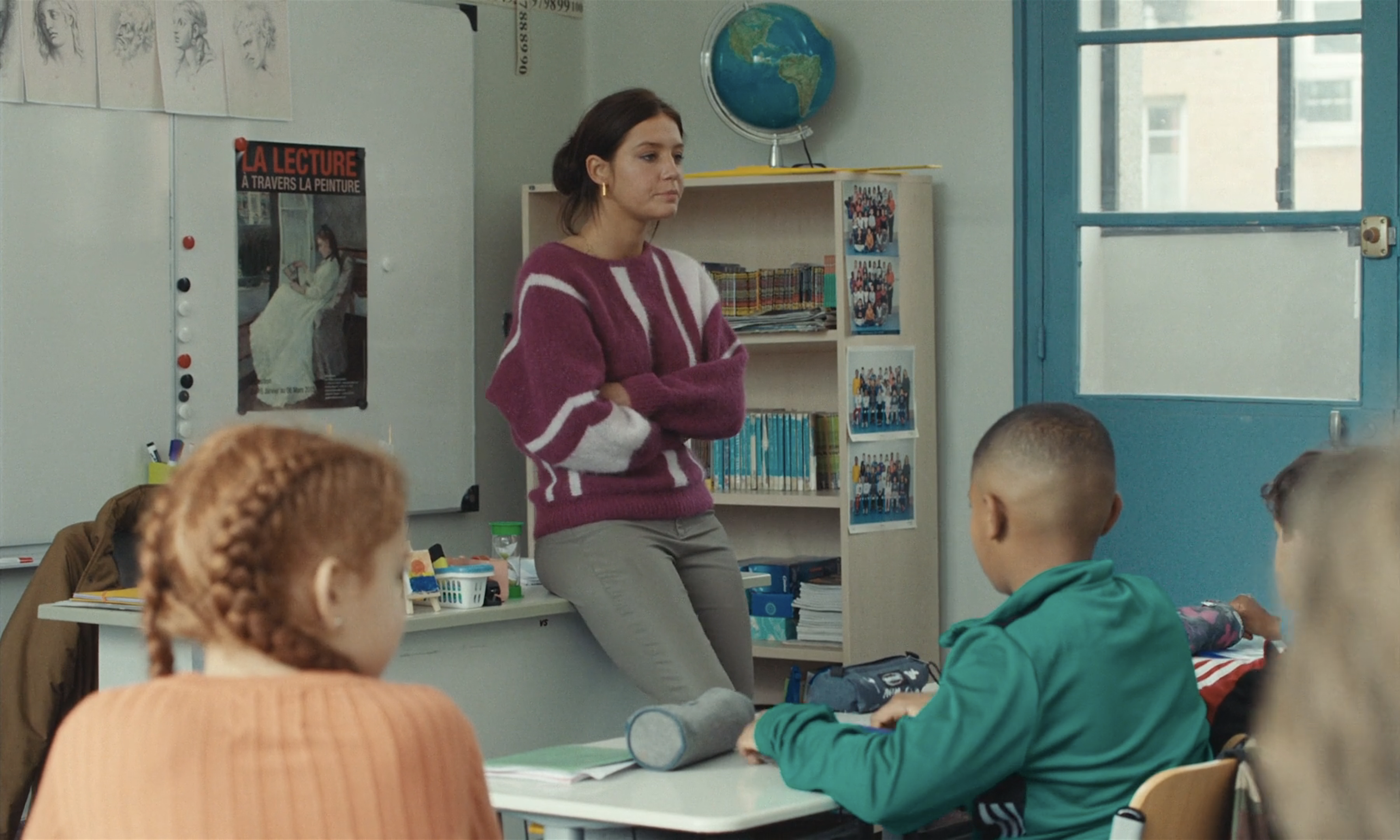 Adèle Exarchopolous in tight light pants and a purple and white sweater leans against a desk in a classroom as children look on.