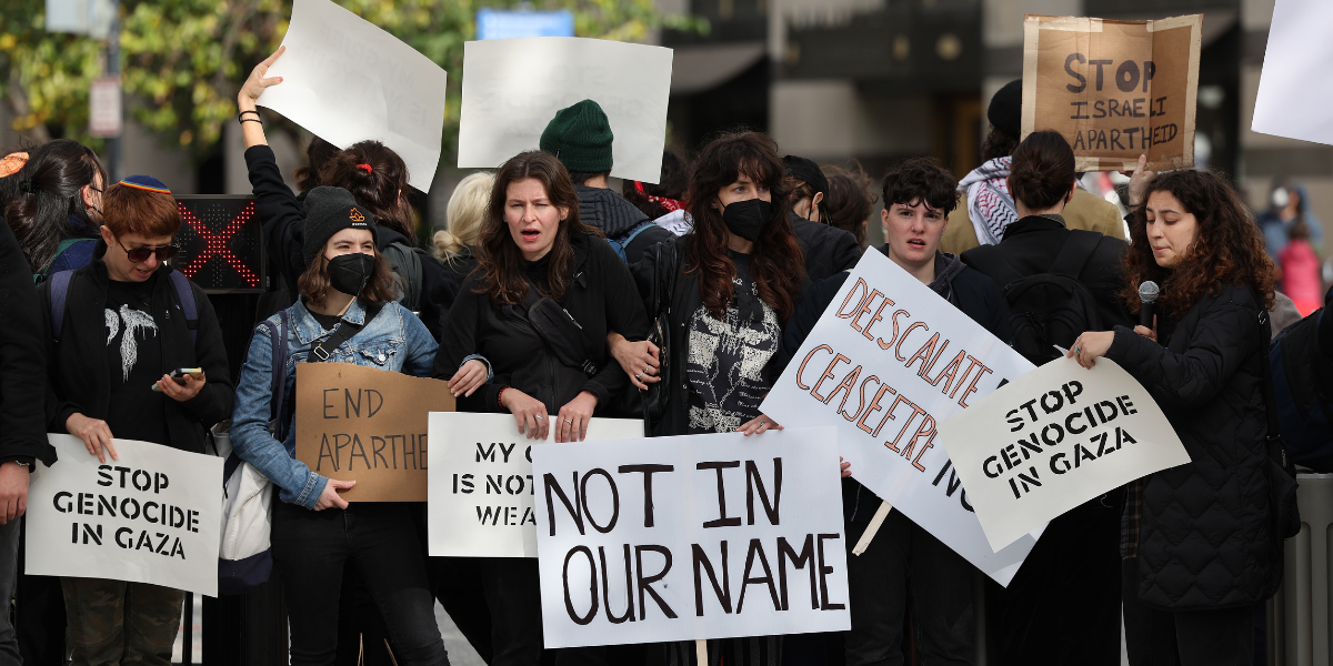 Members of the Jewish Voice for Peace and the IfNotNow movement protest and block access to Pennsylvania Ave. near the White House during a demonstration to support Gaza on October 16, 2023 in Washington, DC. Signs read: NOT IN OUR NAME. END APARTHEID. STOP THE GENOCIDE. DE-ESCALATE / CEASEFIRE.