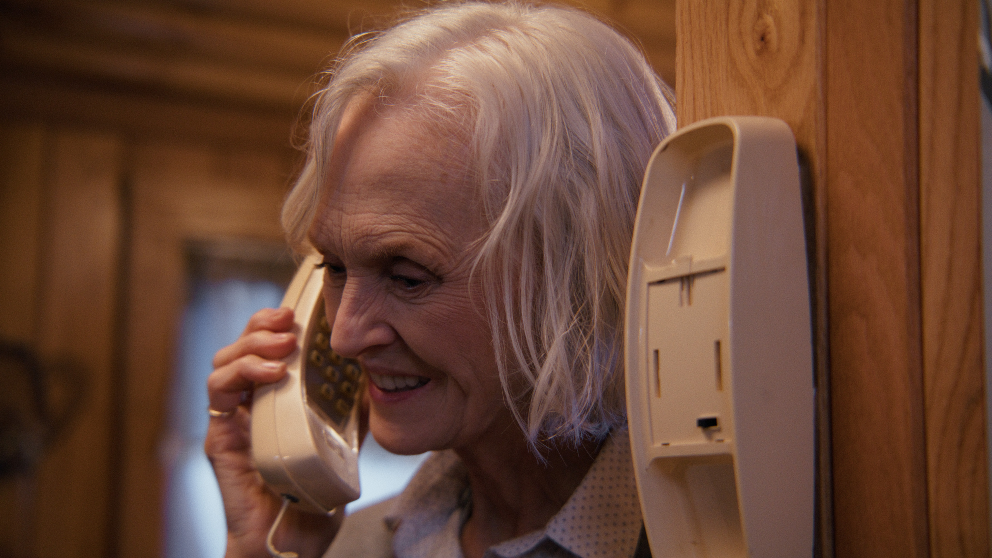 A old woman talks on a landline while leaning against a wooden wall.