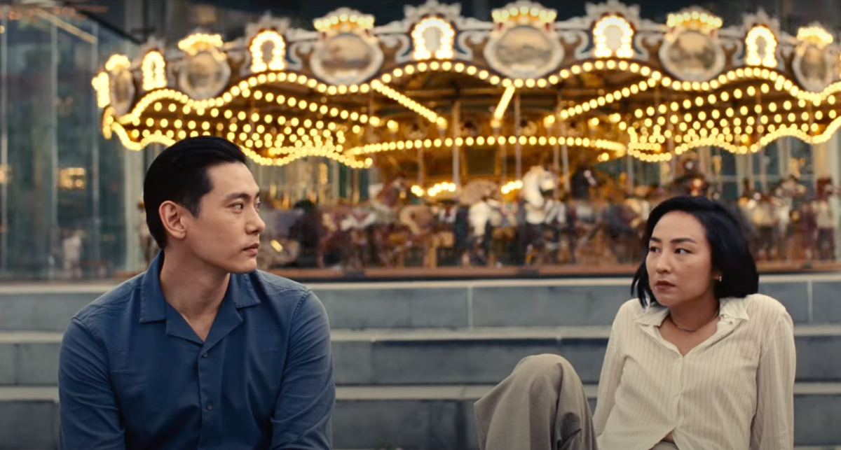 Greta Lee and Teo Yoo sit on stairs in front of a carousel and look at one another.