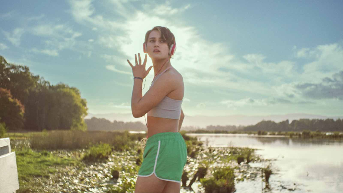 A trans teenage girl dances with headphones on, a beautiful lake behind her.