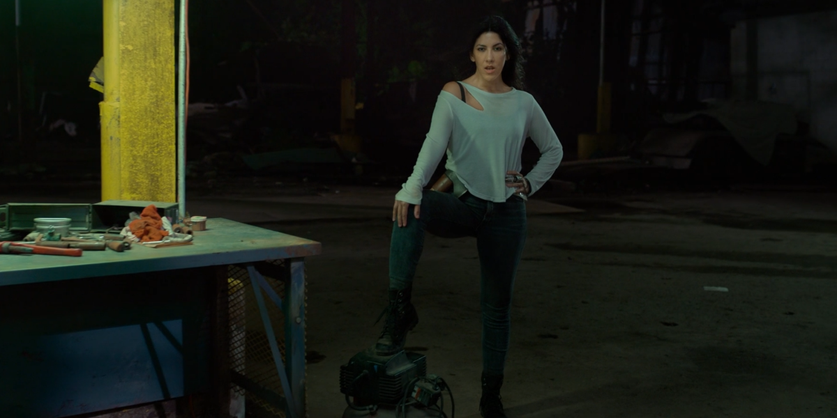 Stephanie Beatrix in a loose blue top and tight black pants lifts one leg up onto a car engine.