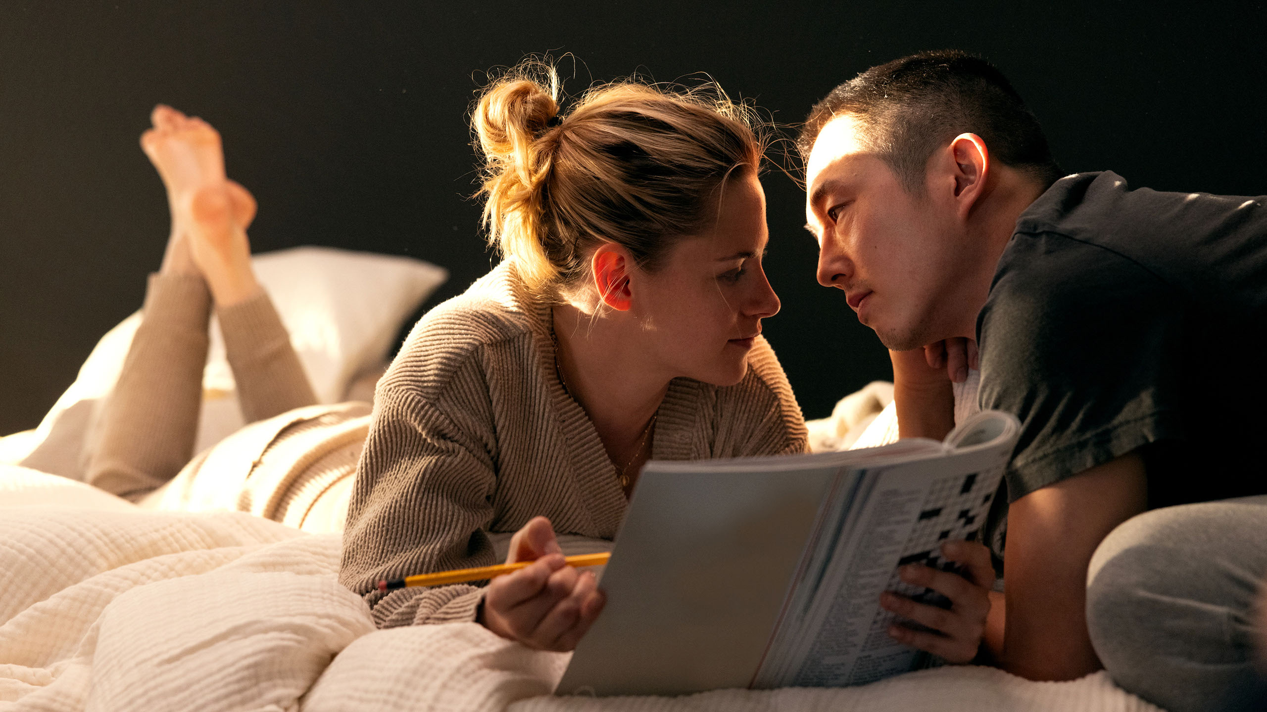 Kristen Stewart lies on a bed reading a book and leans in to kiss Steven Yeun.