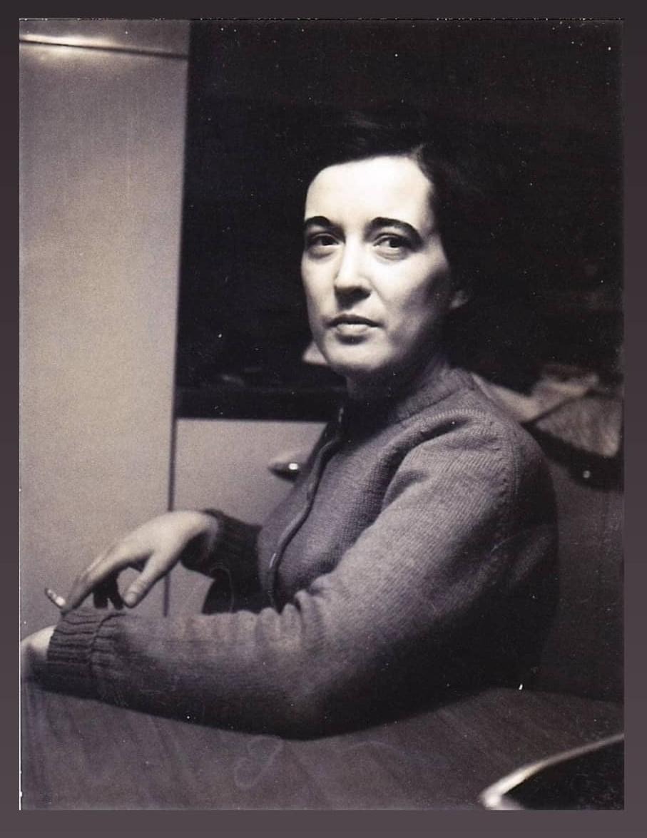 a black and white photo of nico's grandma, looking like she is in her mid to late 30's, with dark short hair and severe shadows crossing her face. she is wearing a sweater and smoking a cigarette, sitting at a table, torso turned away from the viewer but face turned and regarding the camera. her expression is subtle, a slight smile with her eyes, mysterious