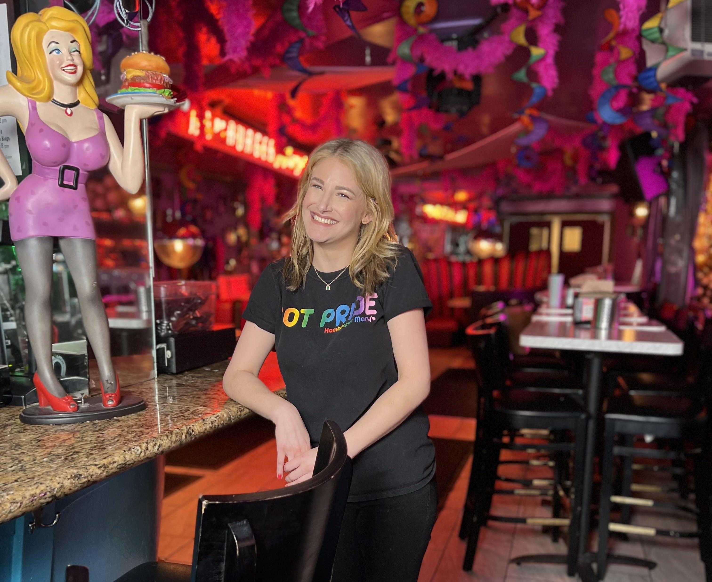 Cheyanne Dunn smiles, leaning on the bar at Hamburger Mary's next to a smiling Hamburger Mary's statue.