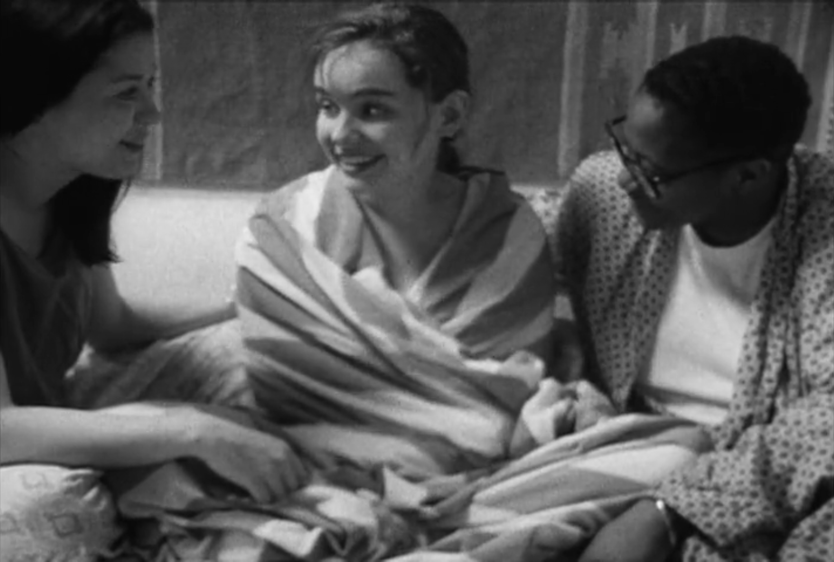 Guinevere Turner sits on a couch wrapped in a blanket with two lesbians on either side of her.